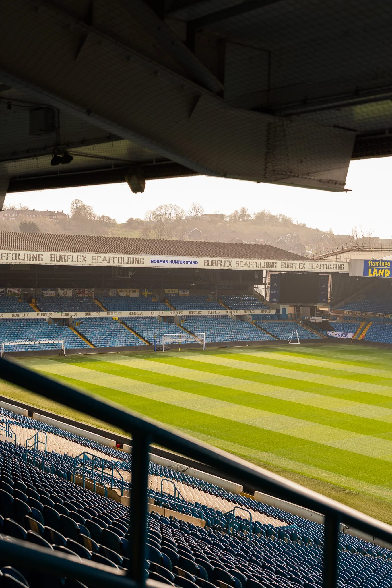 Empty soccer stadium with green and yellow striped grass field, blue seats, and advertising banners.