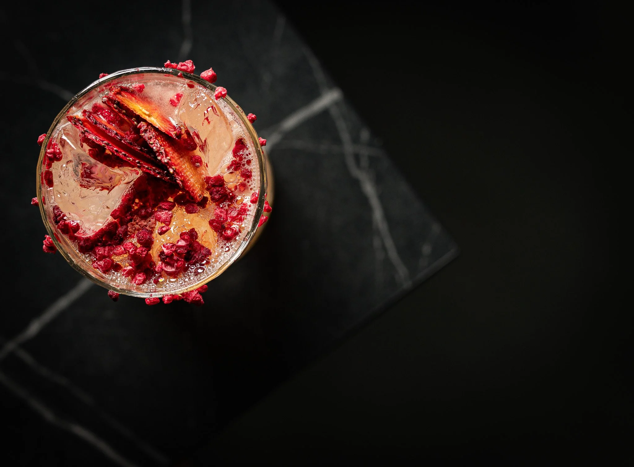 Top view of a cocktail glass with ice, red garnish, and a dried citrus peel on a black surface.