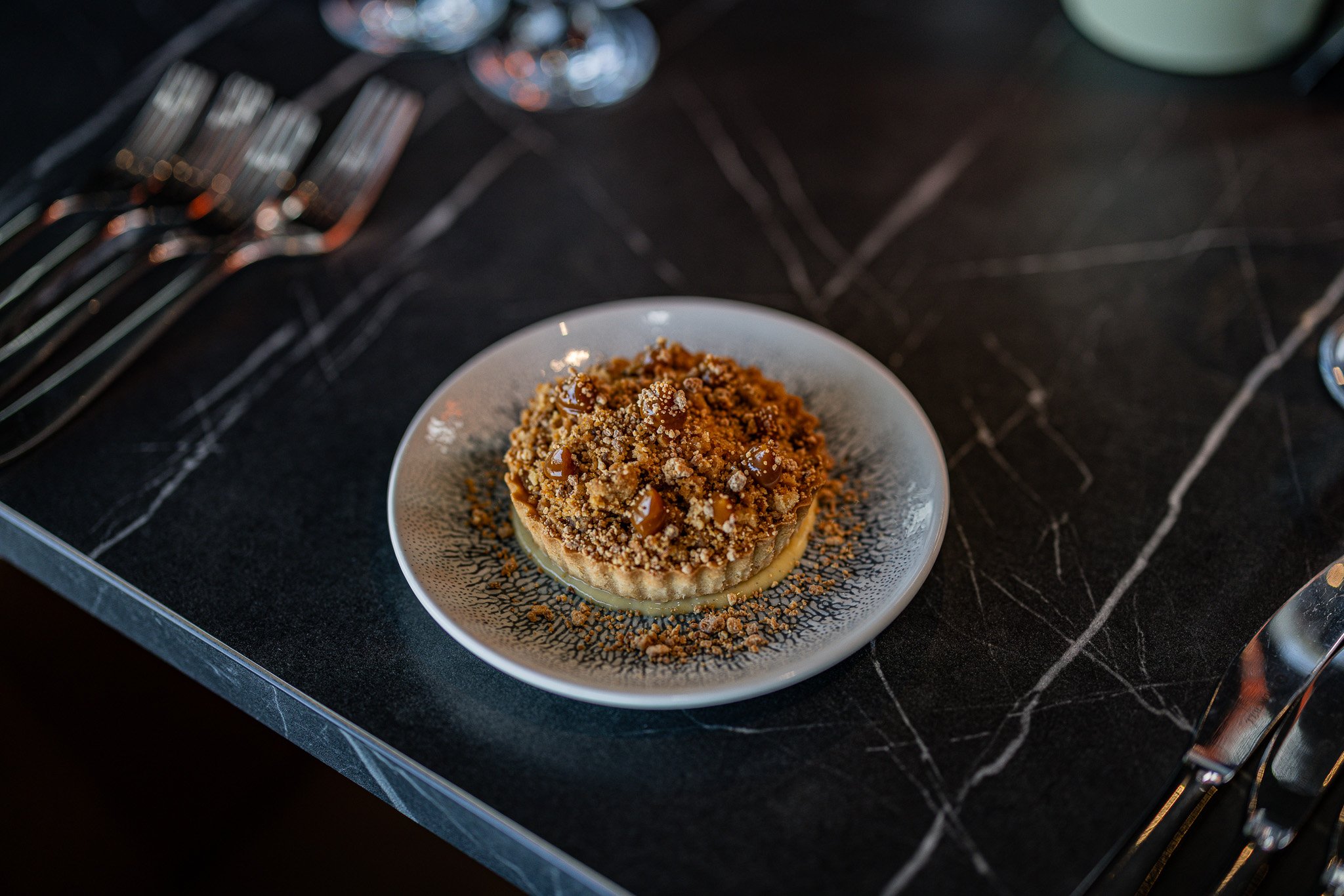 A dessert topped with crumbled cookies and caramel sauce on a decorative plate, placed on a black marble table with silverware nearby.