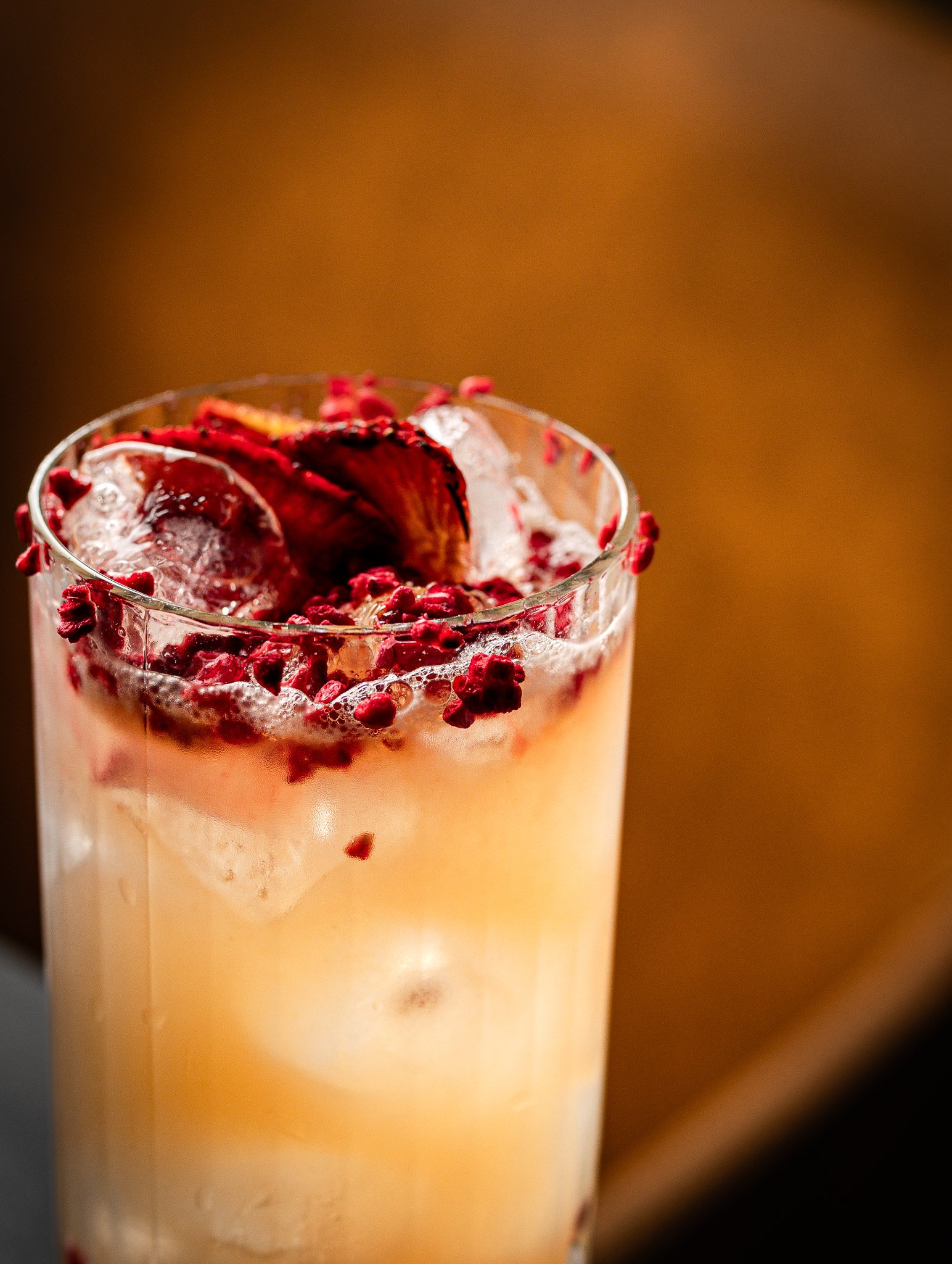 Close-up of a tall glass drink topped with berry garnishes and ice cubes, against a warm blurred background.