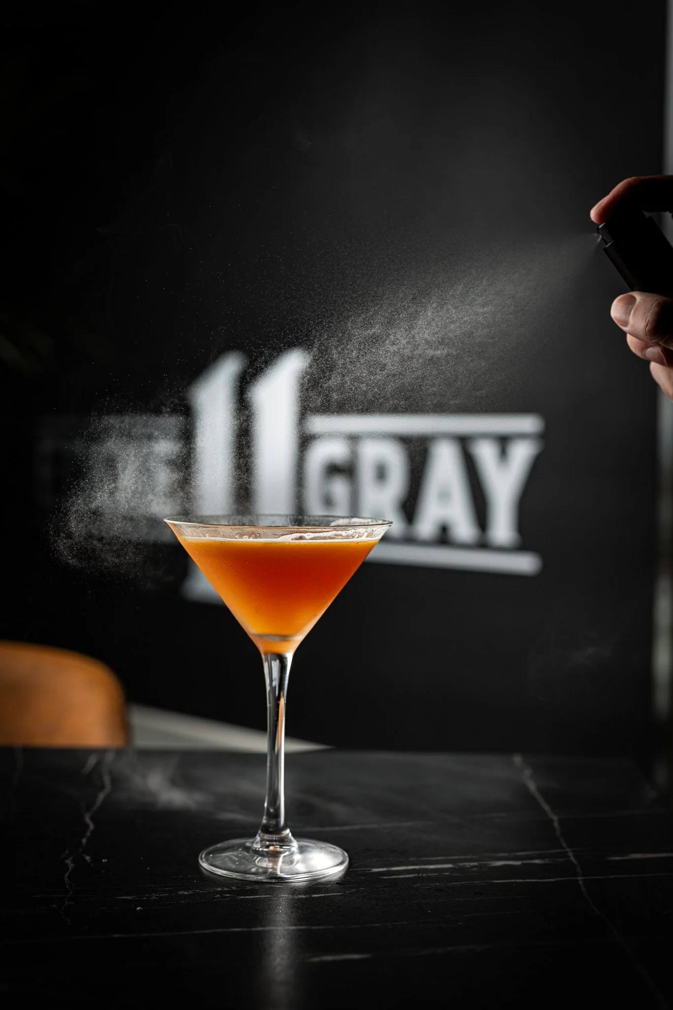 A hand spraying powder onto a peach-colored cocktail in a martini glass with a blackboard background displaying partially visible white text.