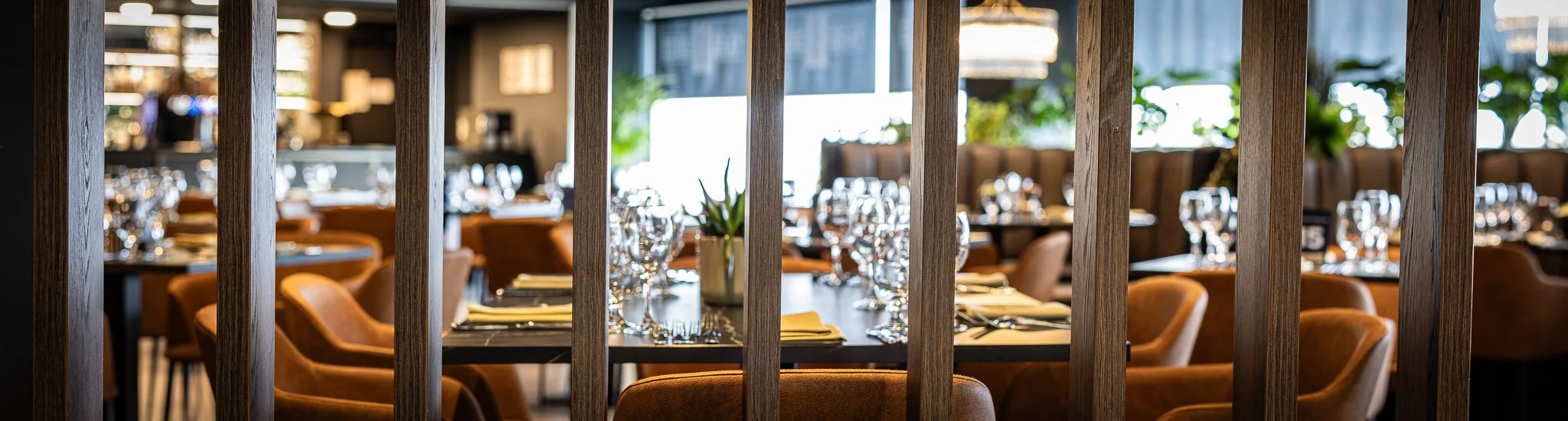 A view of a restaurant dining area seen through wooden vertical slats, with tables set with glassware, plates, napkins, and a potted plant, with more tables, chairs, and windows in the background.