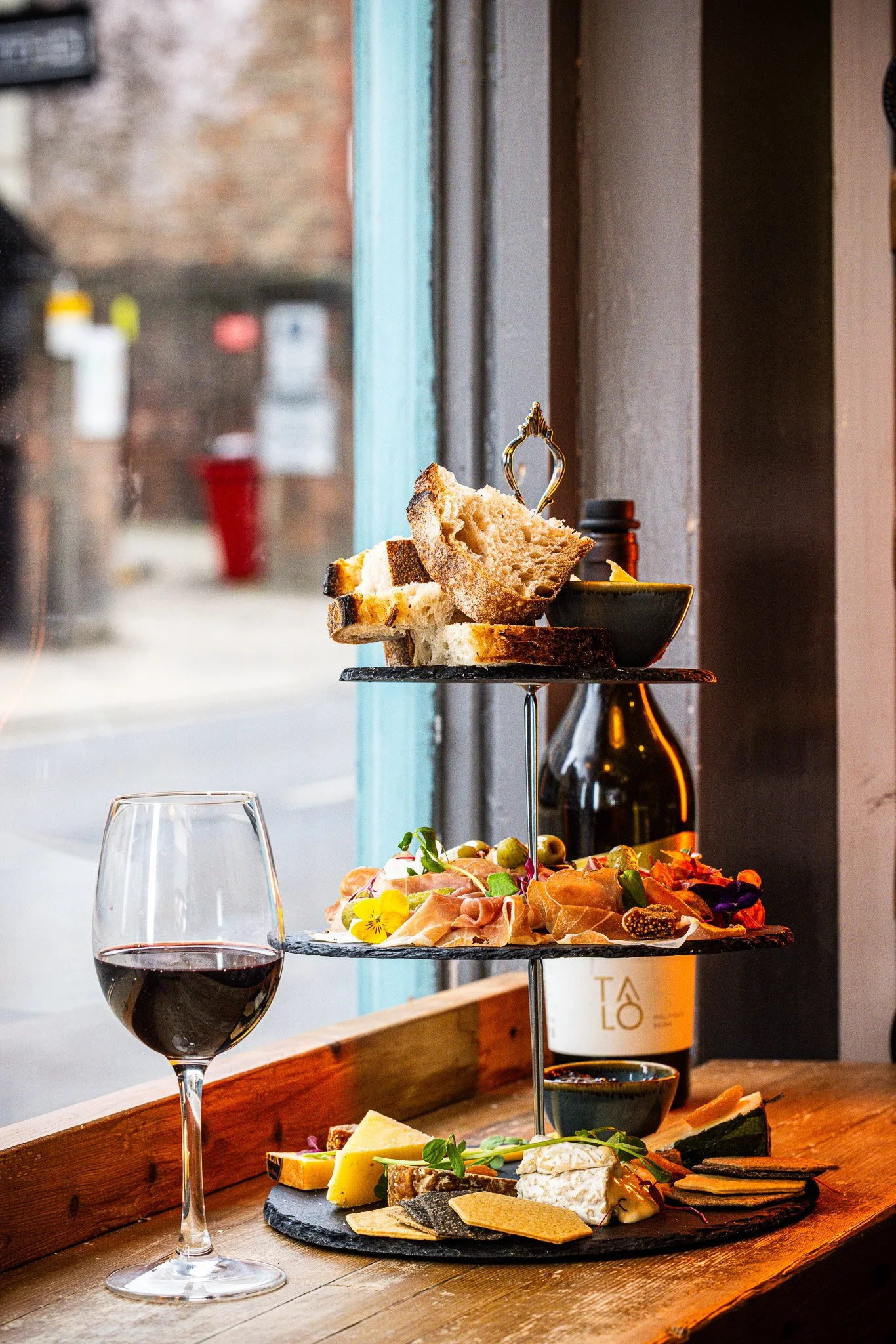 Three-tiered serving tray with bread, cheese, meats, olives, and crackers, next to a glass of red wine on a wooden table near a window.