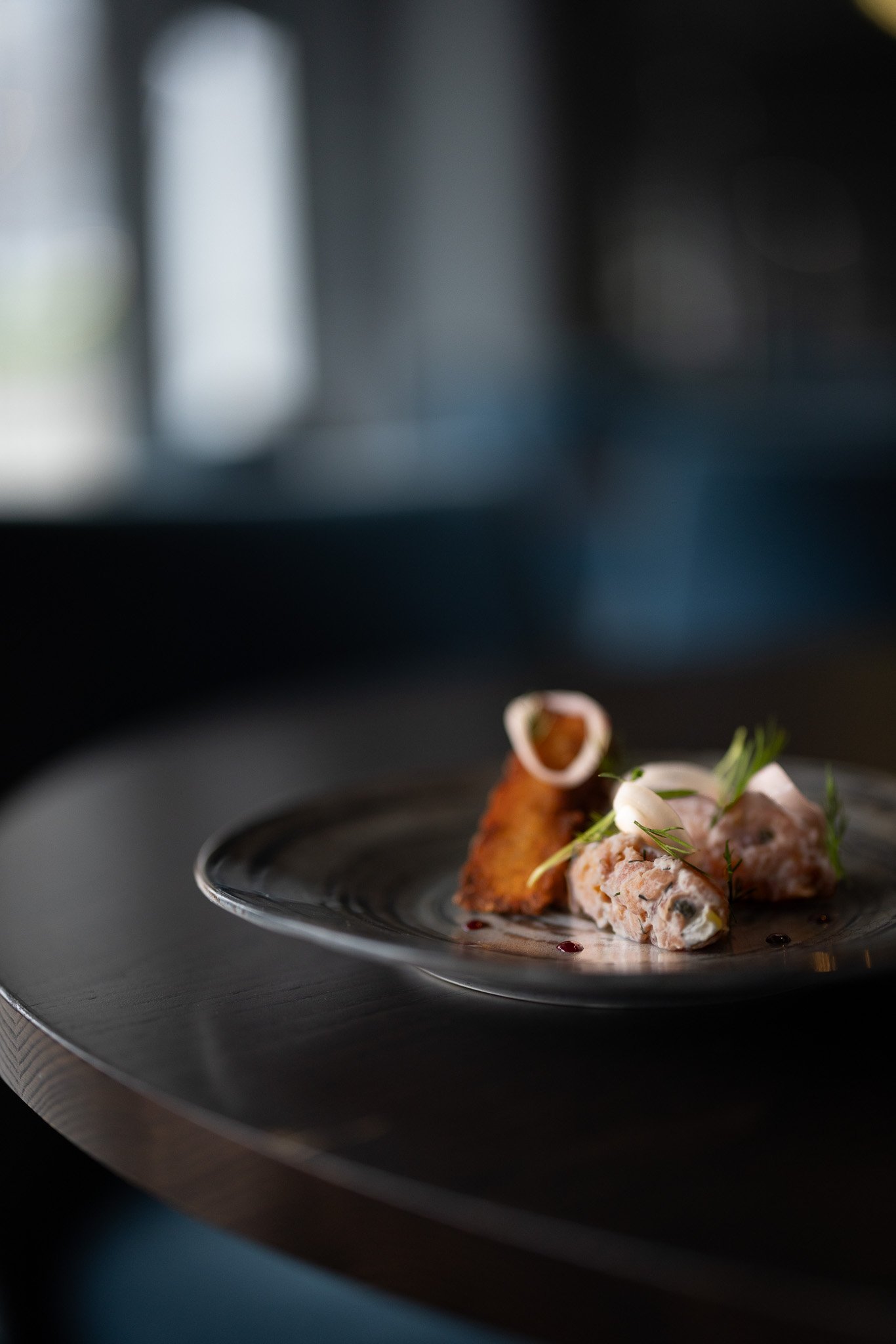 Elegant dish with meat and garnish on a black plate, placed on a wooden surface, with a blurred dark background.