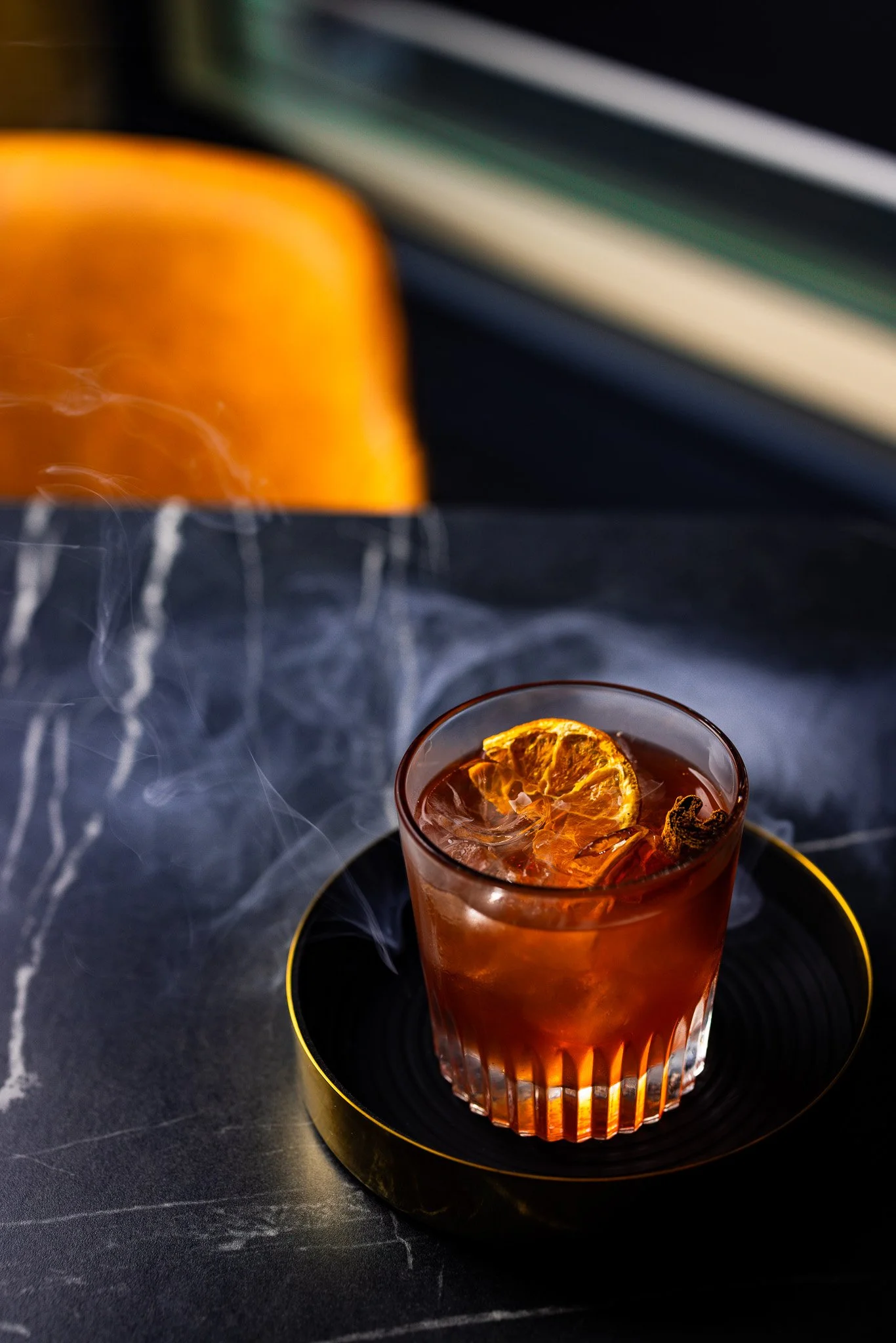 A glass of dark amber beverage with orange slices and ice, placed on a black round tray on a dark marble surface, with a blurred orange object and window view in the background.