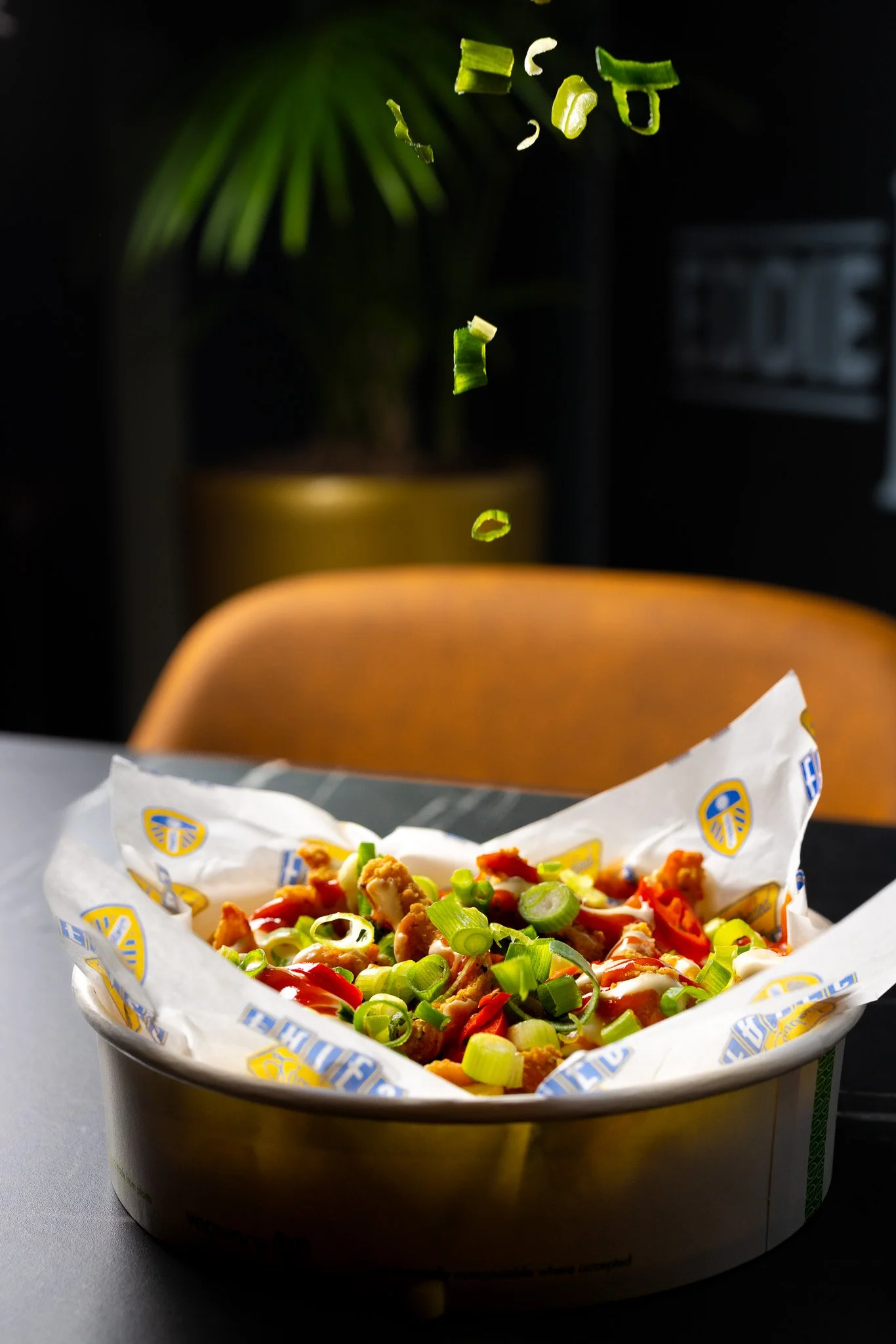 Falling chopped green onions over a bowl of fried chicken and vegetables, on a table with a brown chair and a potted plant in the background.