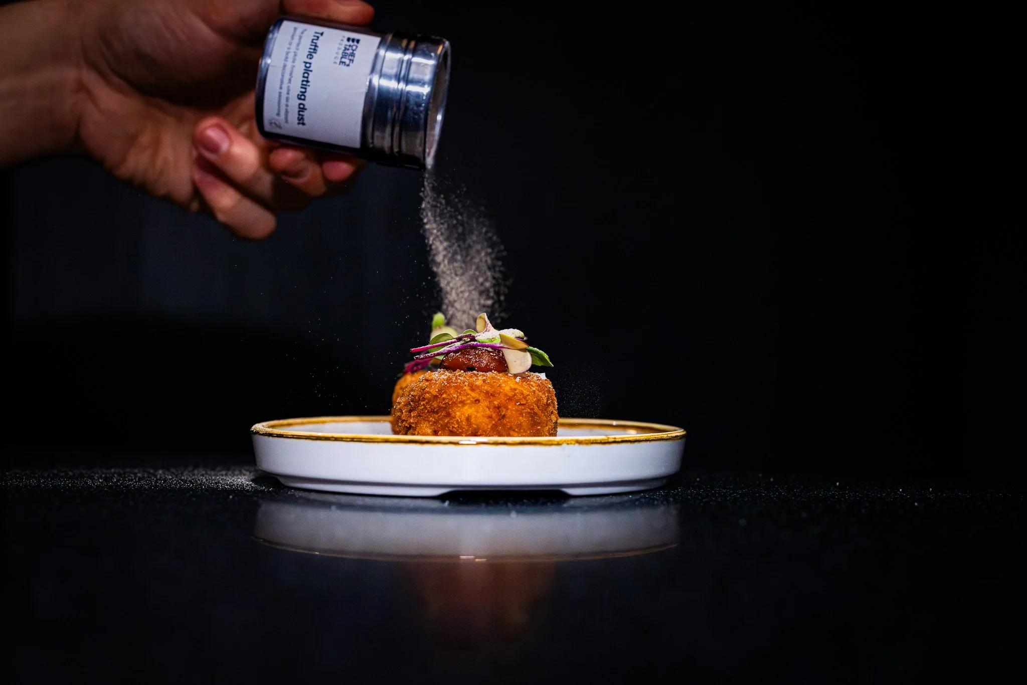 A hand sprinkling seasoning over a gourmet dish on a white plate against a black background.