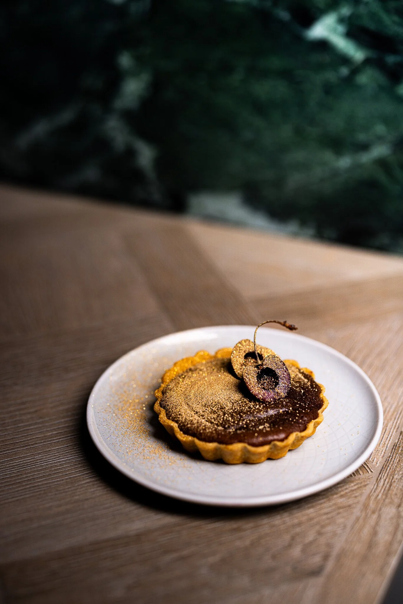 Chocolate tart topped with gold dust and garnished with two cherries with stems, served on a white plate on a wooden surface.