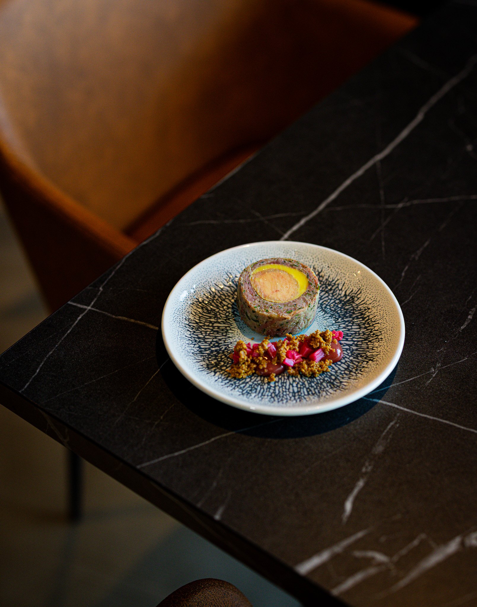 A plated gourmet dish featuring a cylindrical meat or fish roulade with a filling, accompanied by a garnish of crumbled crumbs and pink pickled vegetables on a black and white patterned plate.