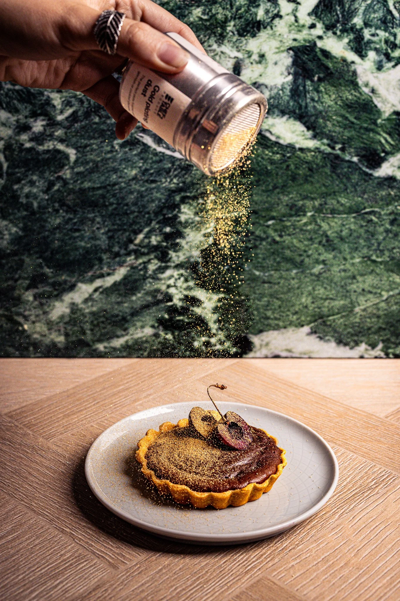 A person sprinkling gold glitter onto a cherry tart on a white plate, with a mountain landscape background in the image.
