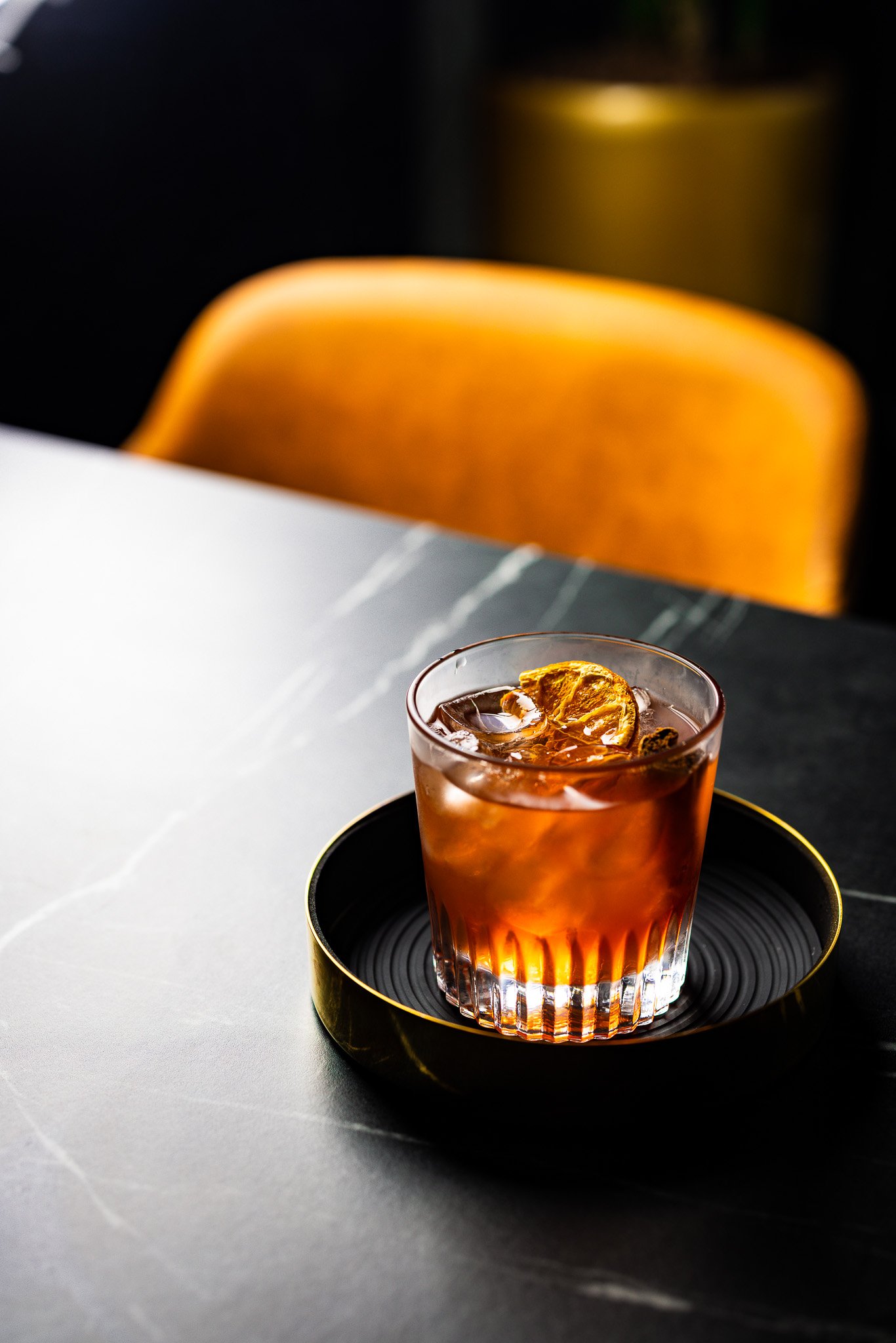 A glass of cocktail with ice and a lemon slice on top, placed on a black tray on a dark gray marble table.