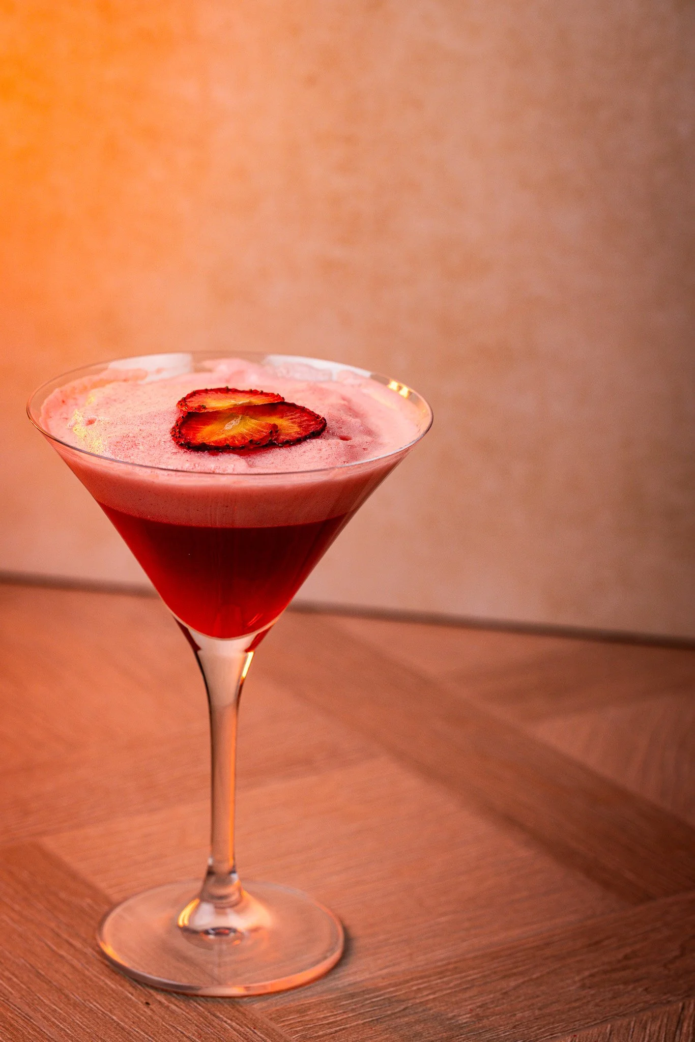 A red cocktail in a martini glass garnished with strawberry slices on top, placed on a wooden surface.