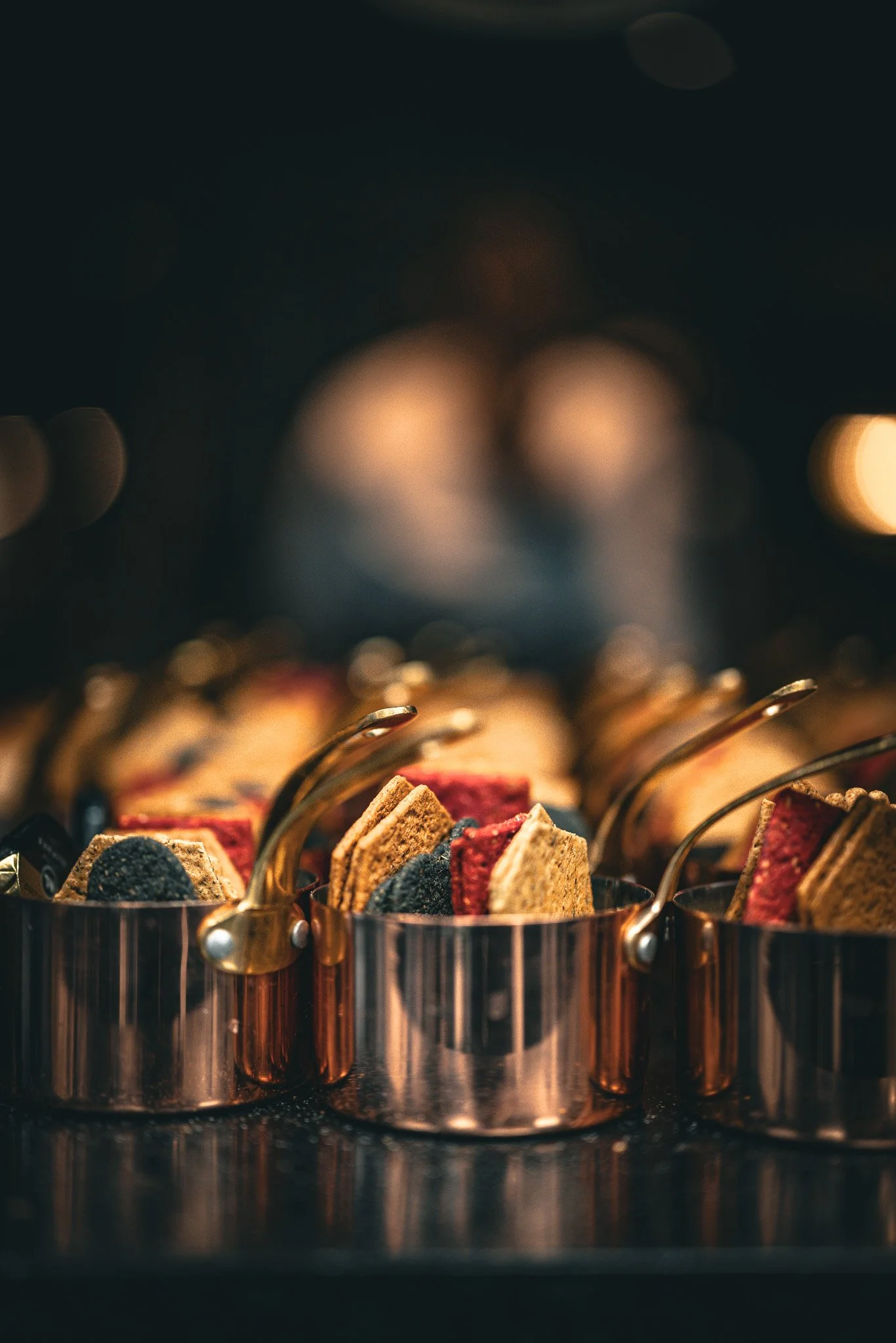 Close-up of assorted cookies in small metallic containers, on a dark surface with blurred lights in the background.