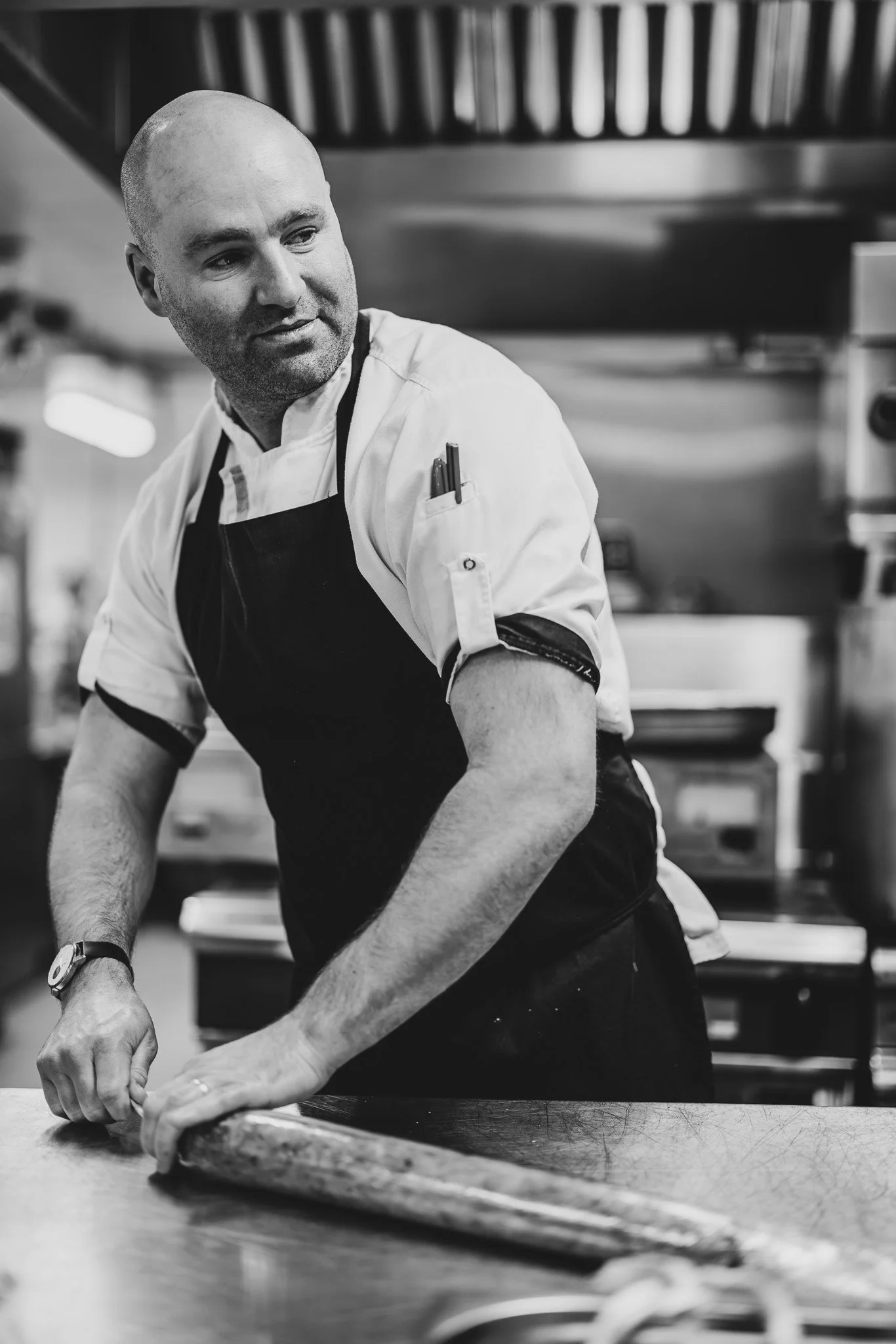 A male chef in a white uniform and black apron prepares fish on a cutting board in a commercial kitchen.