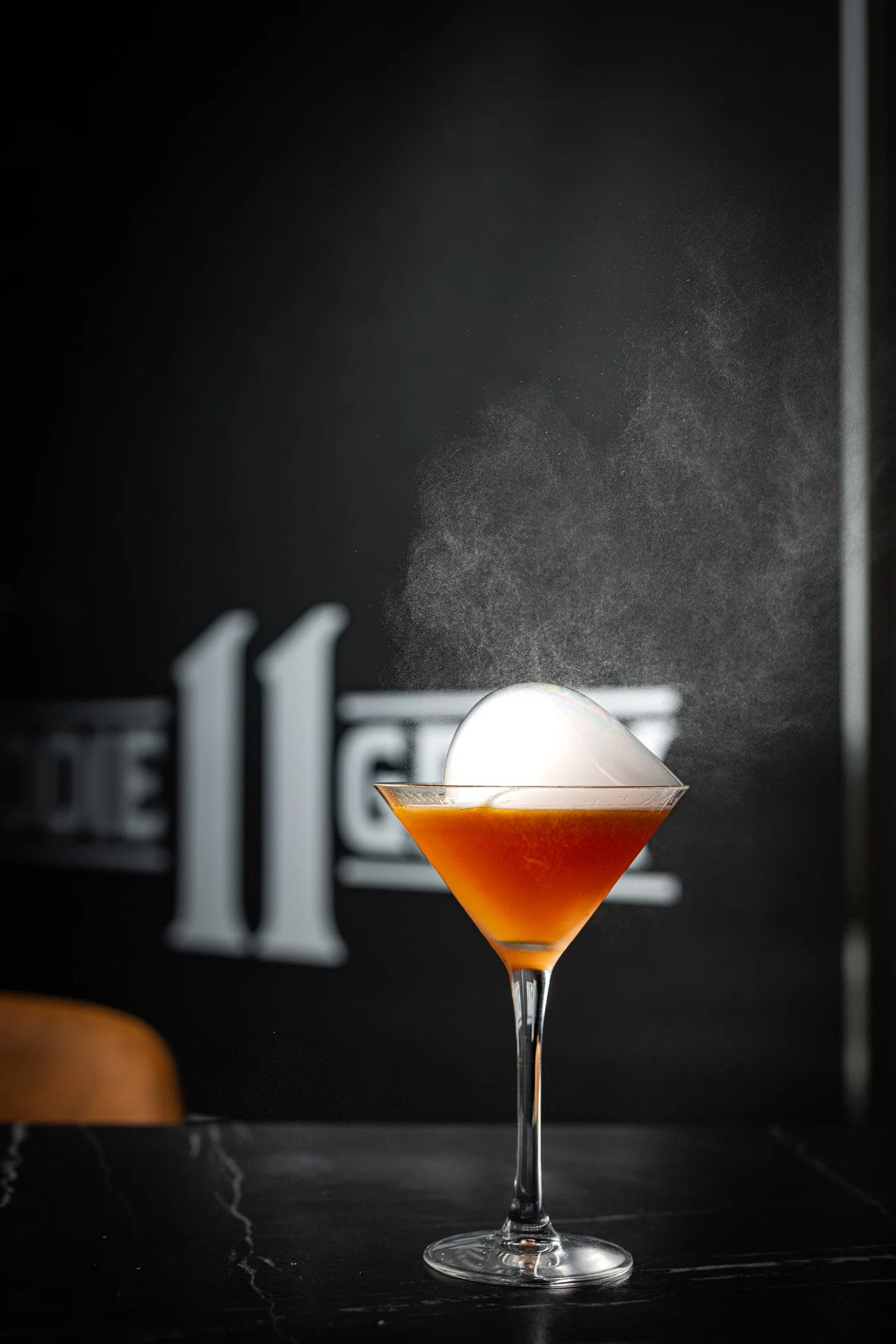 A cocktail in a martini glass with an orange hue and a large ice cube, set against a dark background with a faint logo.