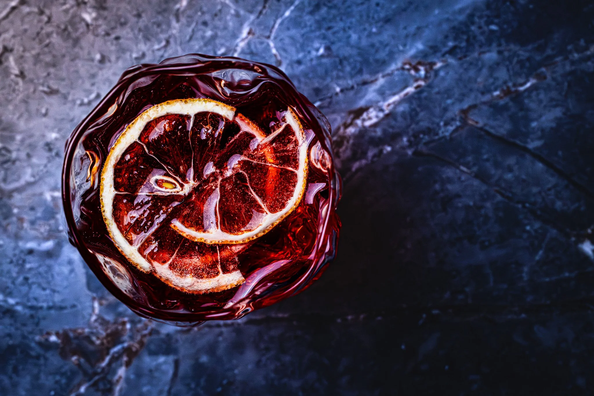 Overhead view of a glass with red beverage and lemon slices, placed on a dark marble surface.