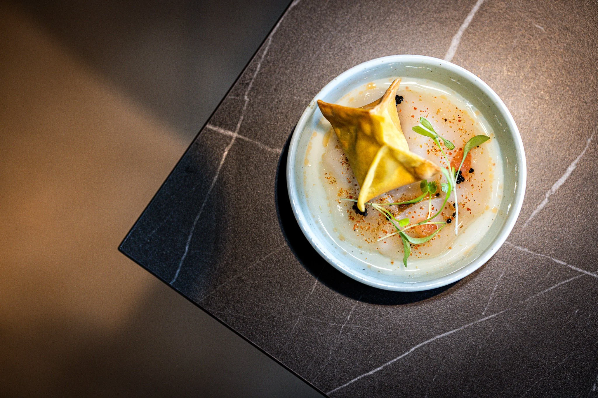 A bowl of soup garnished with microgreens, black pepper, and a crispy wonton chip, placed on a dark marble table.