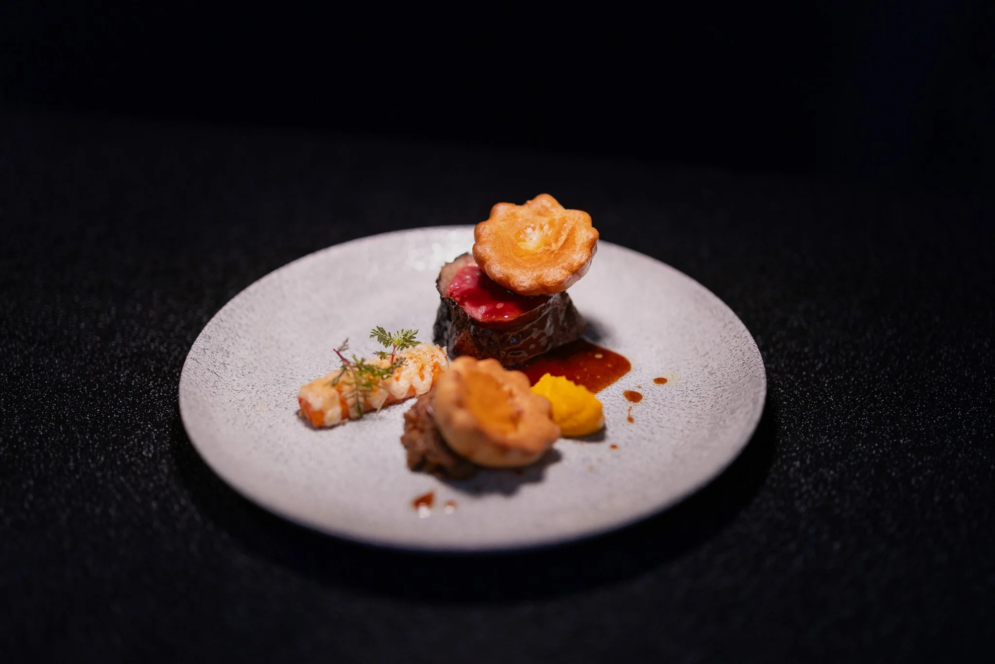Elegant plated gourmet dish with two main meat courses topped with fried garnishes, accompanied by a small piece of lobster, a dollop of yellow sauce, and garnished with a sprig of herbs, served on a white textured plate.