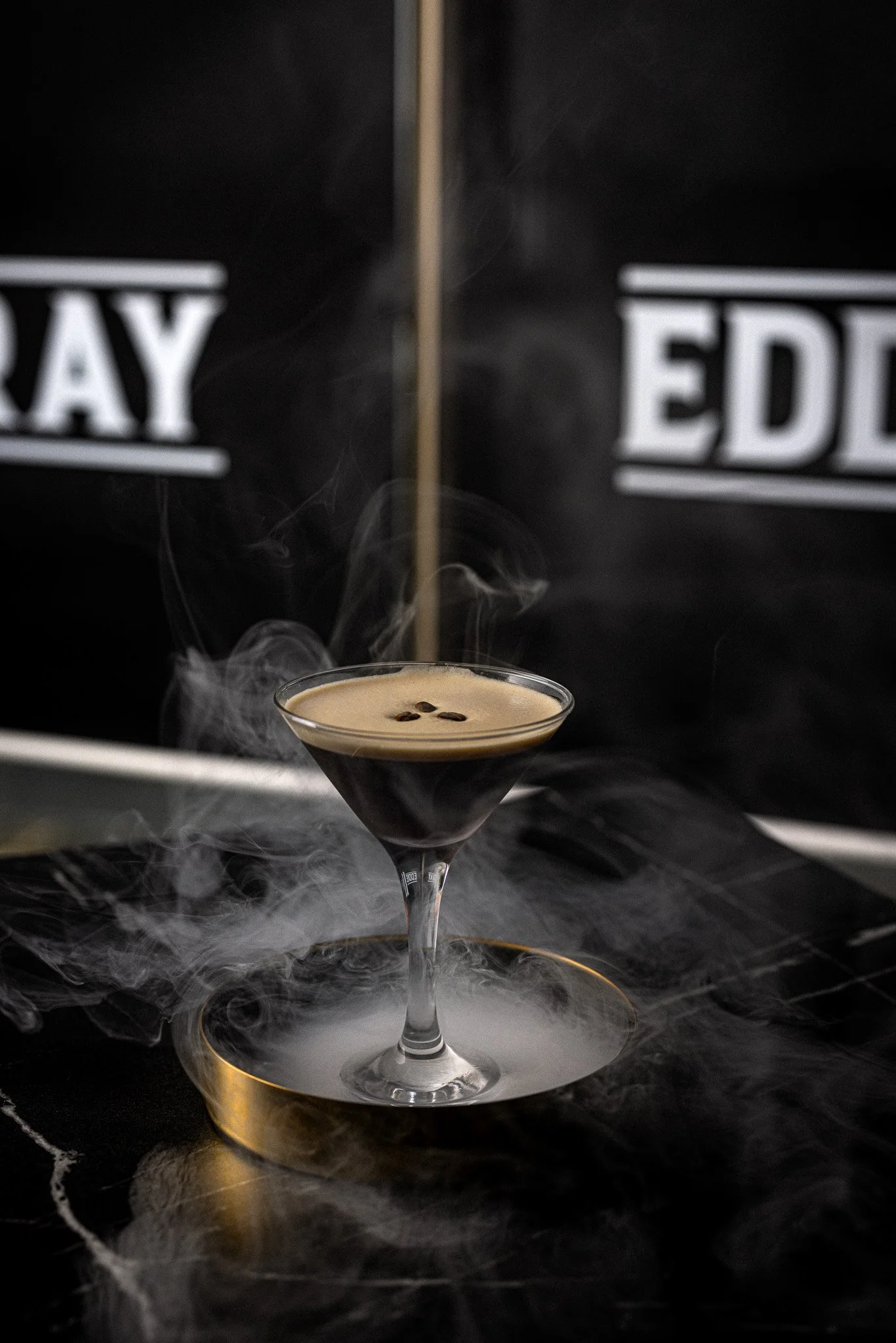 A black coffee cocktail garnished with coffee beans, served in a martini glass surrounded by dry ice smoke, with a dark background featuring partially visible letters.