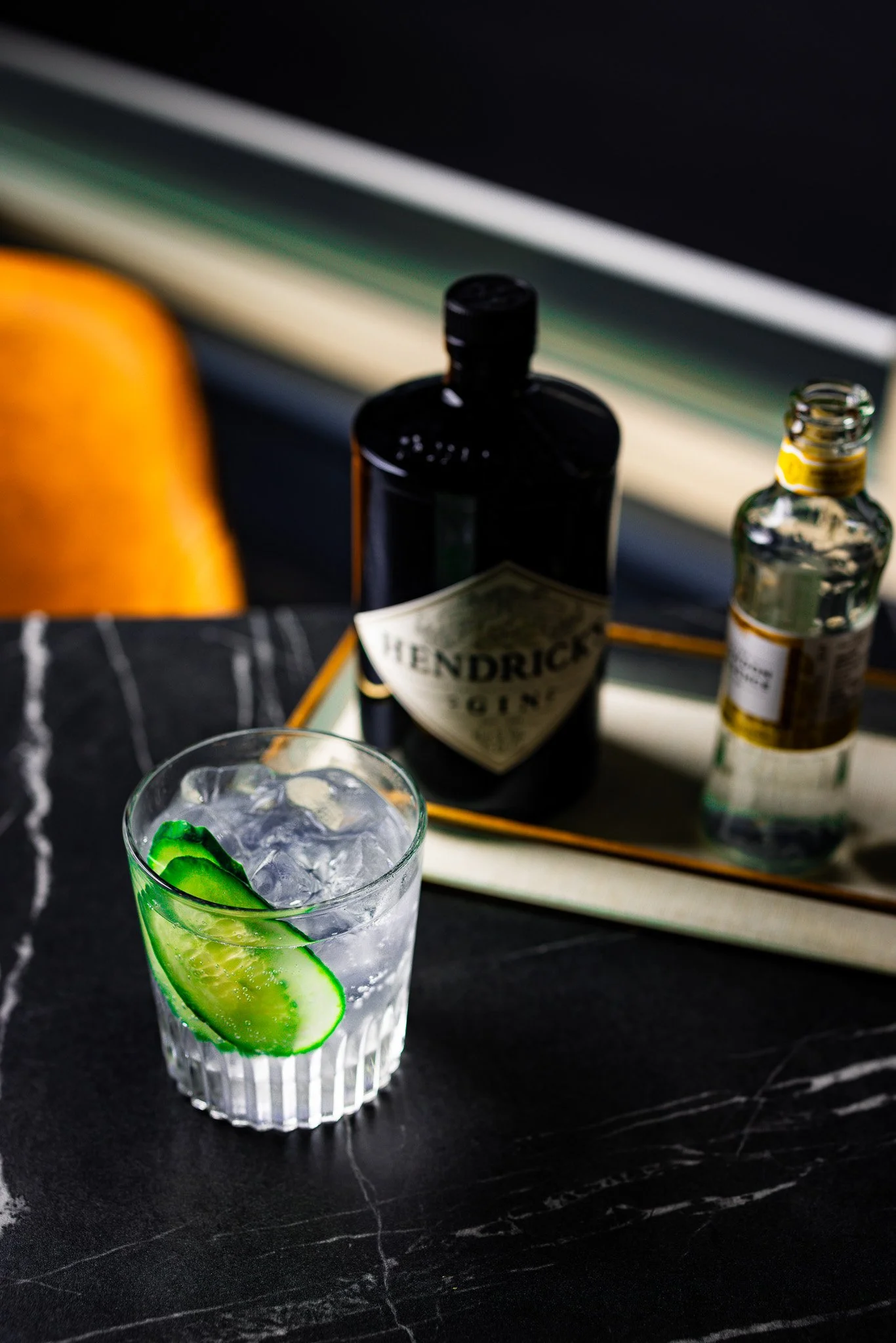 A glass with ice cubes and cucumber slices on a dark marble surface, with bottles of Hendrick's gin and tonic on a tray in the background.