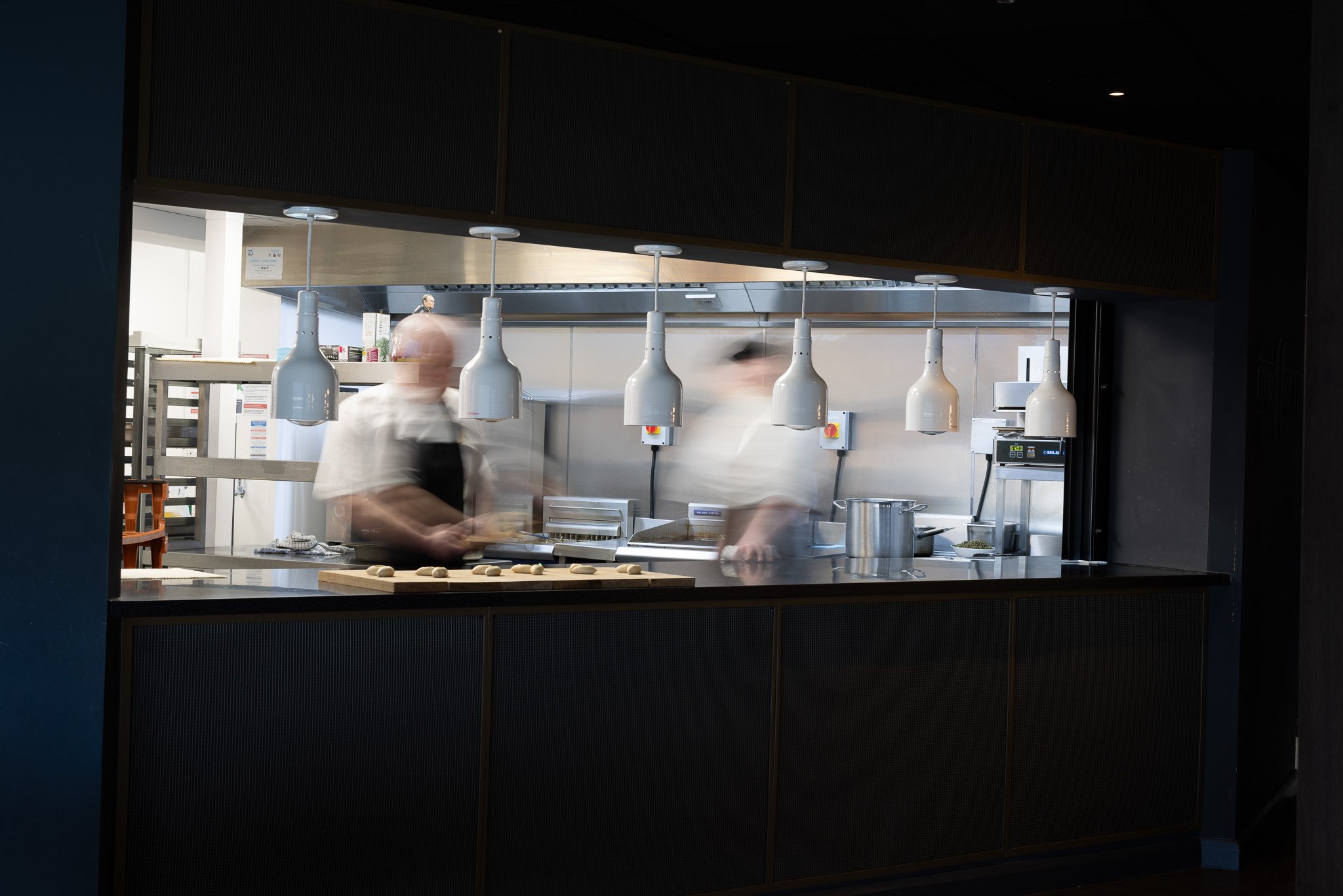 Chef preparing food in a professional kitchen seen through a large open window, with hanging white lamps and stainless steel appliances.