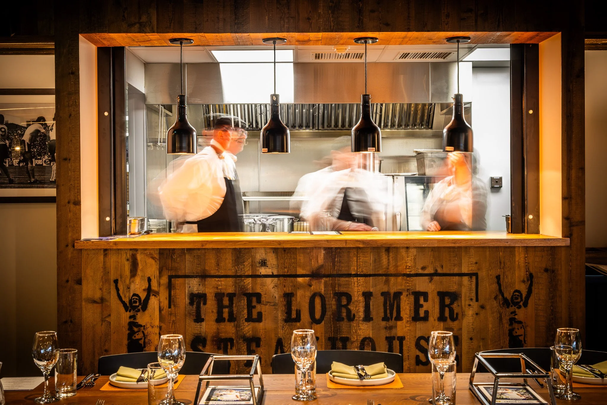 View of a restaurant kitchen seen through a large window with chefs at work, and a wooden bar with artwork and table settings in the foreground.