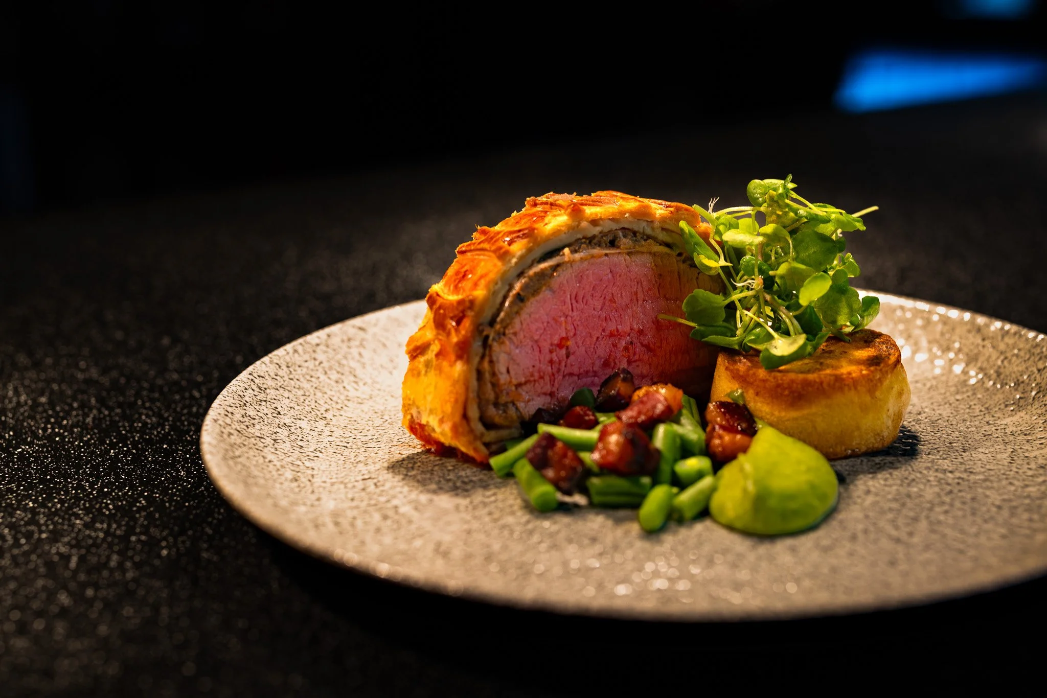 A plated gourmet beef Wellington with a side of microgreens, roasted potato, and diced green vegetables on a textured white plate against a dark background.