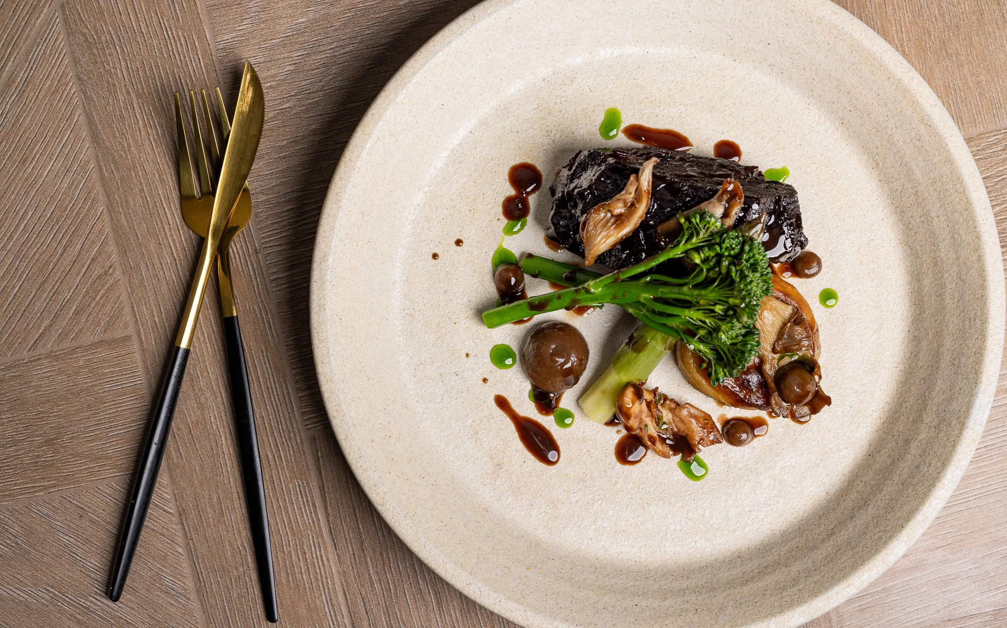 A plate of gourmet food featuring braised meat, broccoli, mushrooms, and sauce on a beige plate, with gold and black utensils on a wooden table.