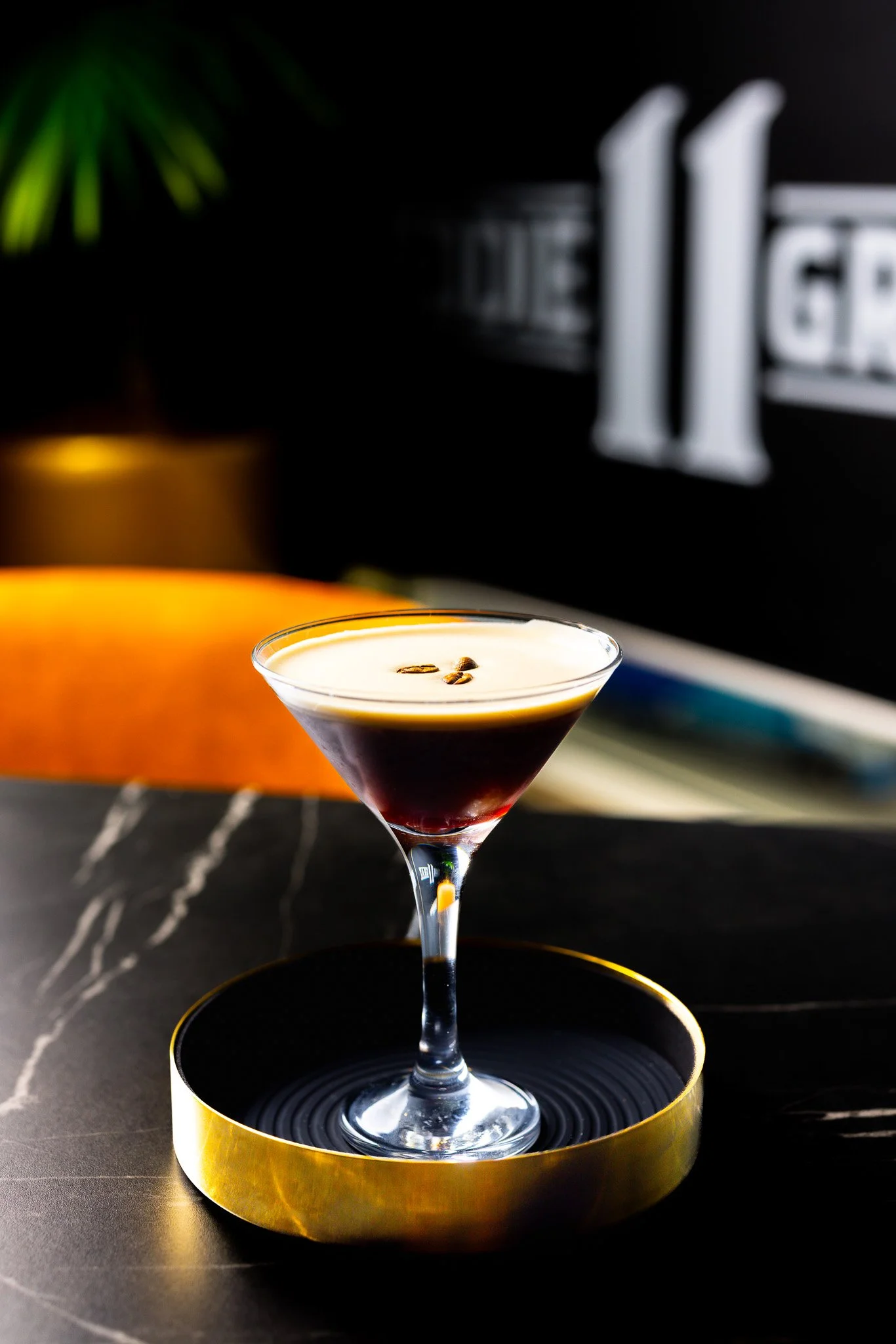 A cocktail in a martini glass with a dark red layer at the bottom, topped with cream and garnished with coffee beans, placed on a round black and gold tray.