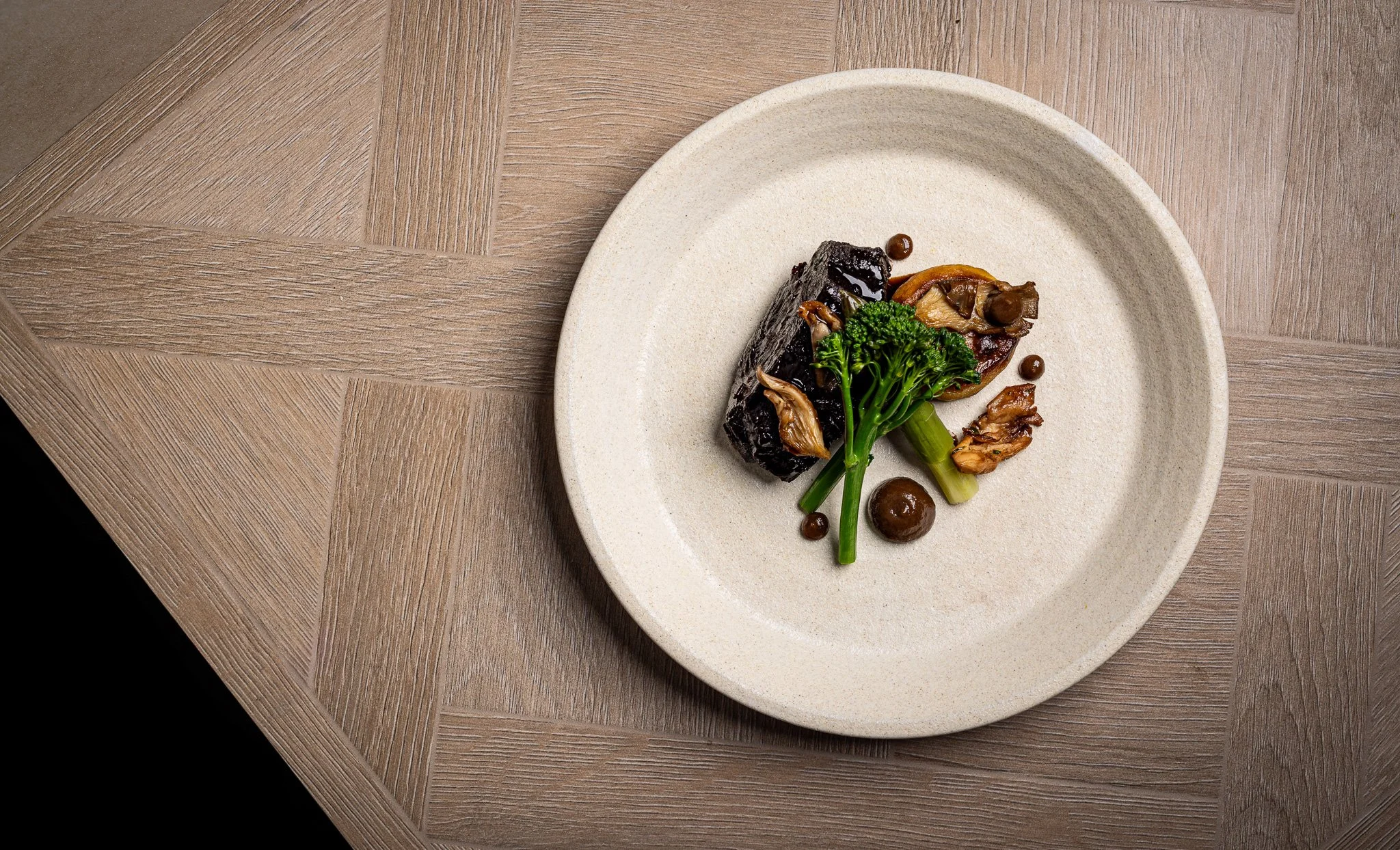 A beige ceramic plate on a wooden table holds roasted vegetables including broccoli, mushrooms, eggplant, and celery, arranged simply.