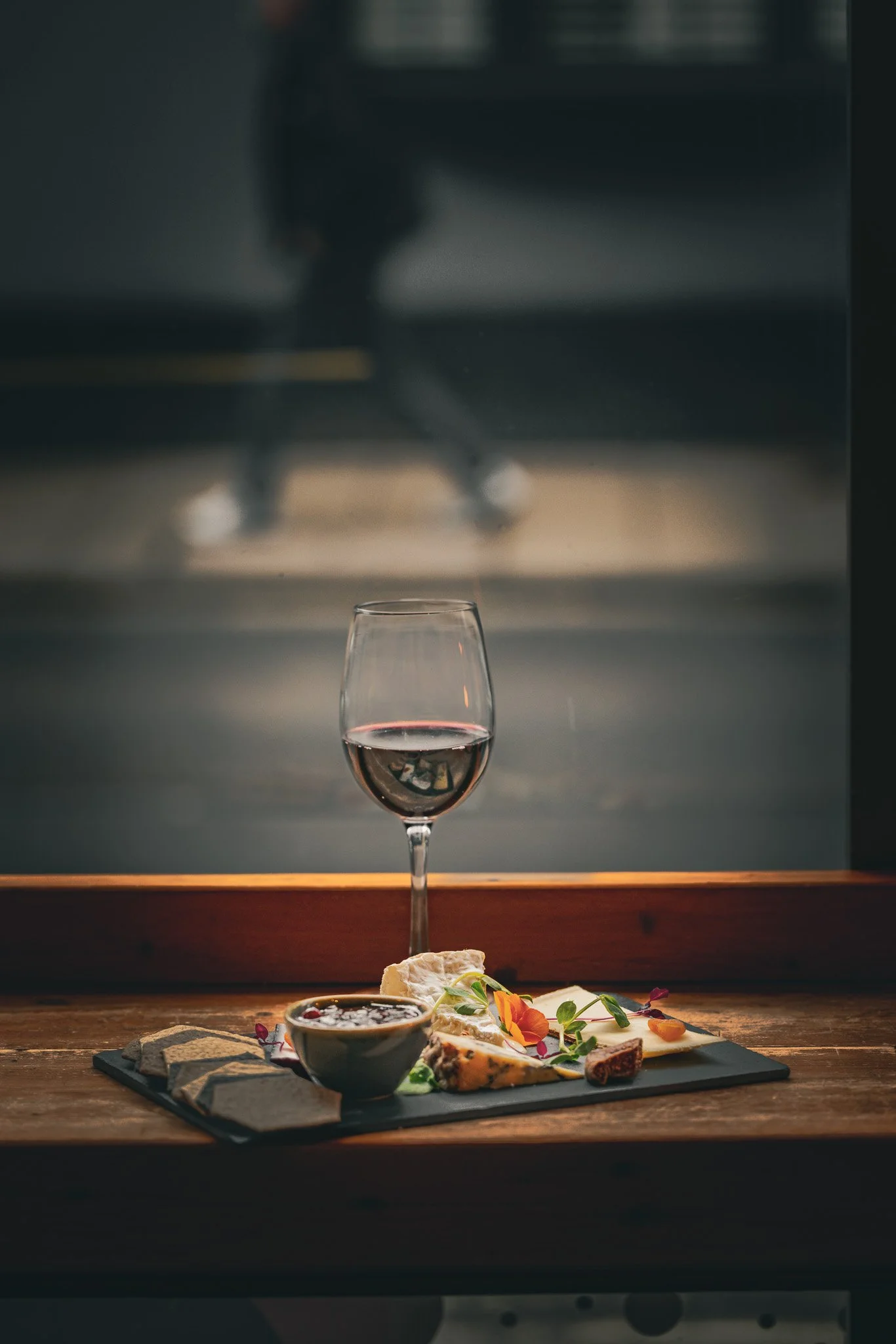 A glass of red wine, a slate cheese platter with assorted cheeses, crackers, jam, and garnishes, on a wooden table near a window with a blurry figure walking outside.