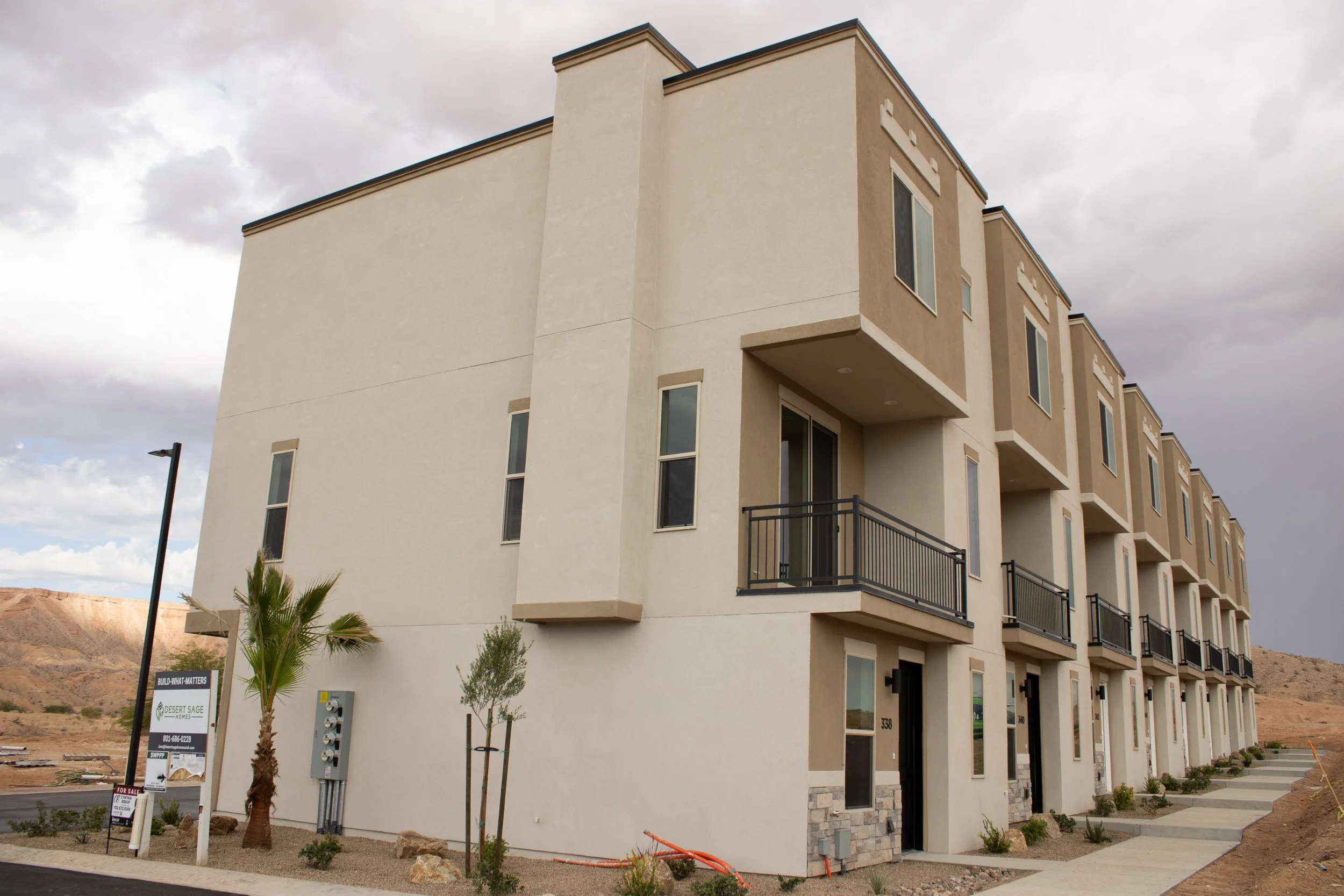 Exterior view of a row of modern townhouses with beige stucco siding, dark garage doors accented with stacked stone veneer, and exposed outdoor HVAC units.