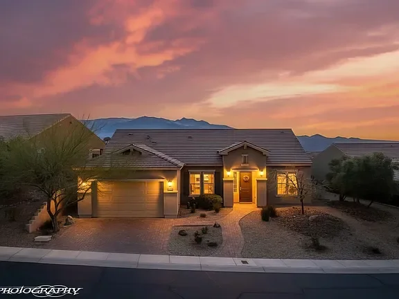 Warm sunset view of a beautifully maintained single-story home in Mesquite, Nevada featuring desert landscaping, glowing exterior lights, and scenic mountain views in the background.
