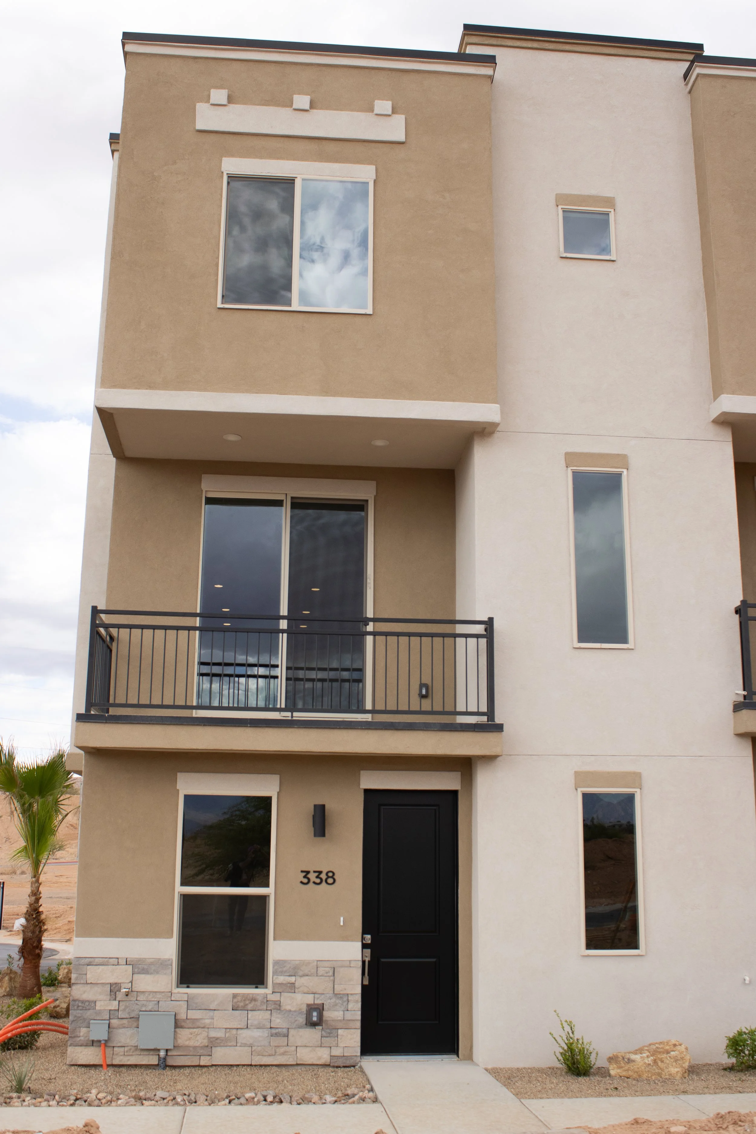 Front exterior of a modern three-story townhouse with beige and white stucco siding, a black front door, a second-floor balcony, and a stacked stone veneer base.