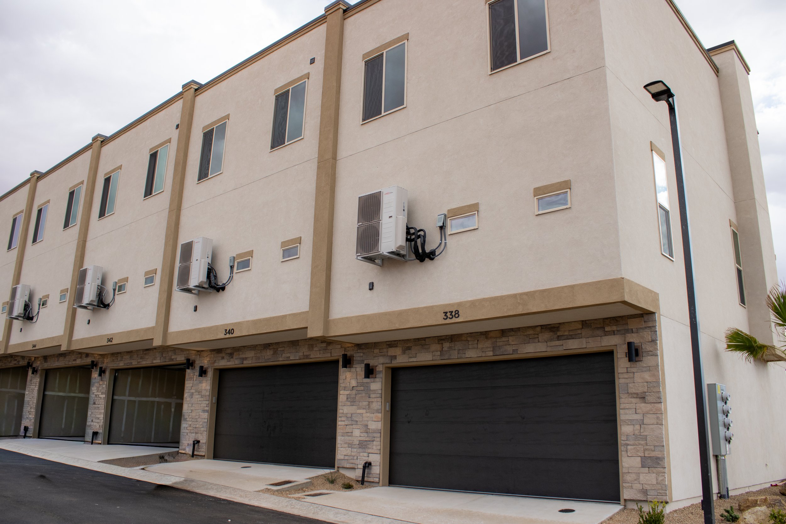 Exterior view of a row of modern townhouses with beige stucco siding, dark garage doors accented with stacked stone veneer, and exposed outdoor HVAC units.