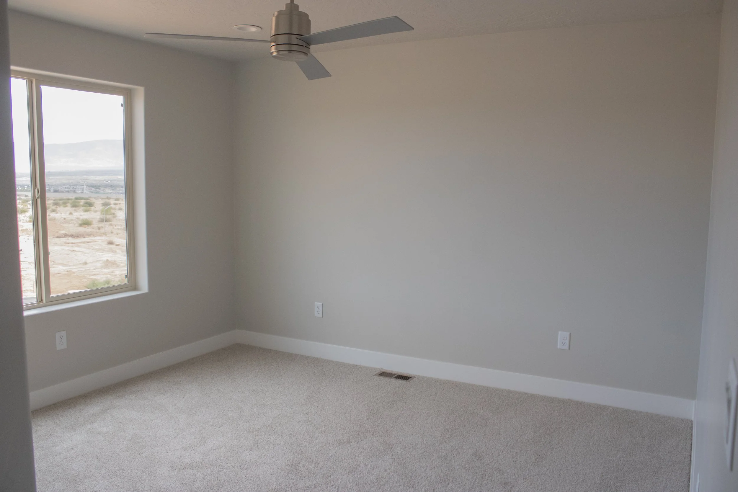 New construction home featuring neutral gray walls, light beige carpet, white baseboard trim, and a modern ceiling fan. A large window offers a view of the surrounding landscape.