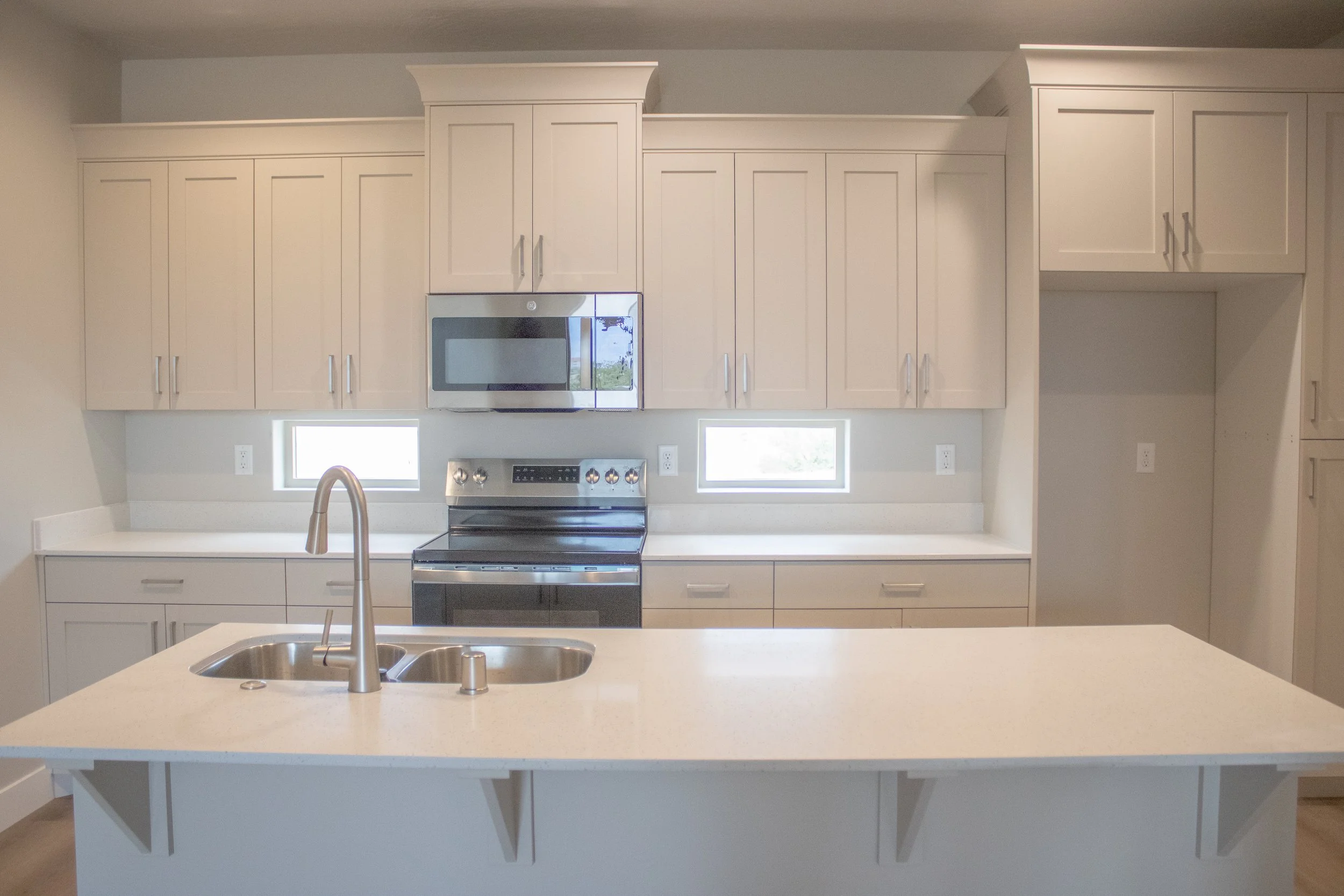 A modern kitchen featuring light beige shaker cabinets, white quartz countertops, and a central island with a double sink. The kitchen includes a black electric range and stainless steel microwave.