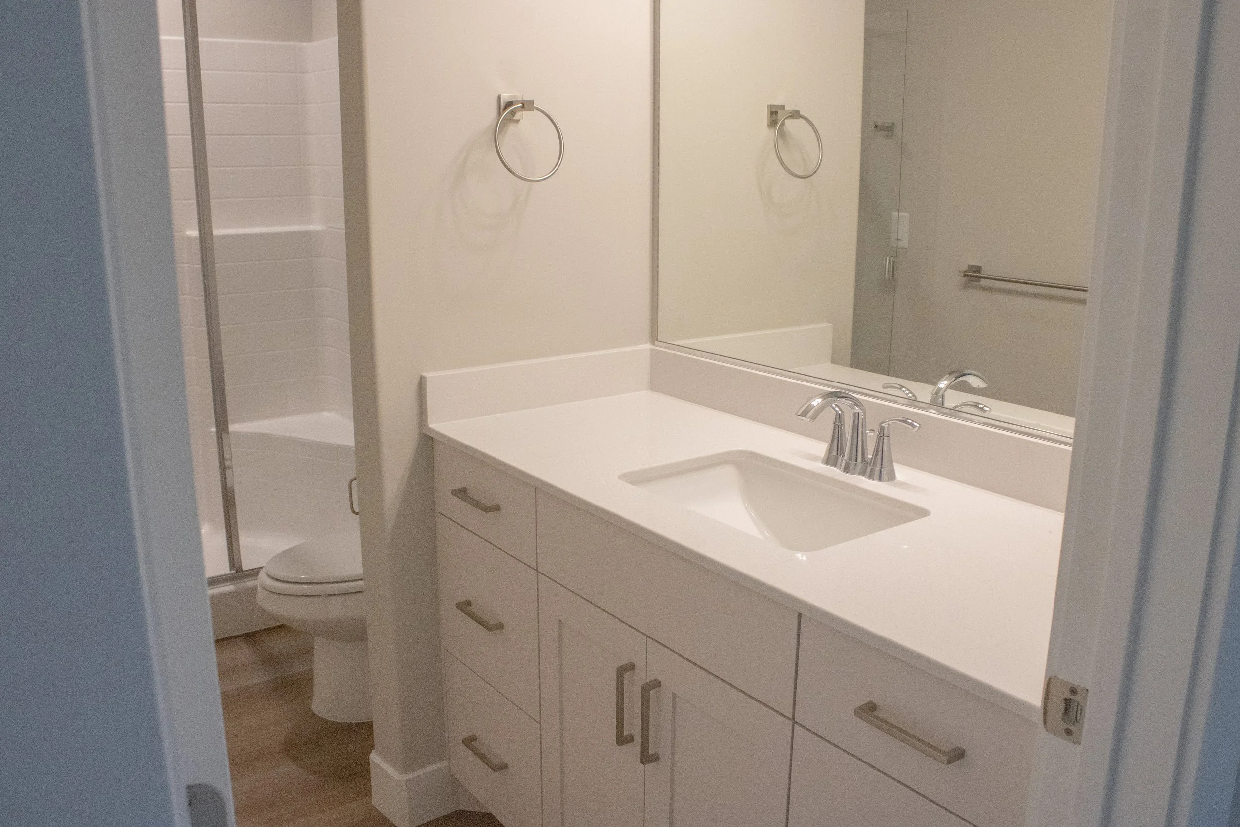 A modern bathroom vanity with white shaker cabinets, a quartz countertop, and an undermount sink. The view looks toward the toilet and glass-enclosed shower with white subway tile.