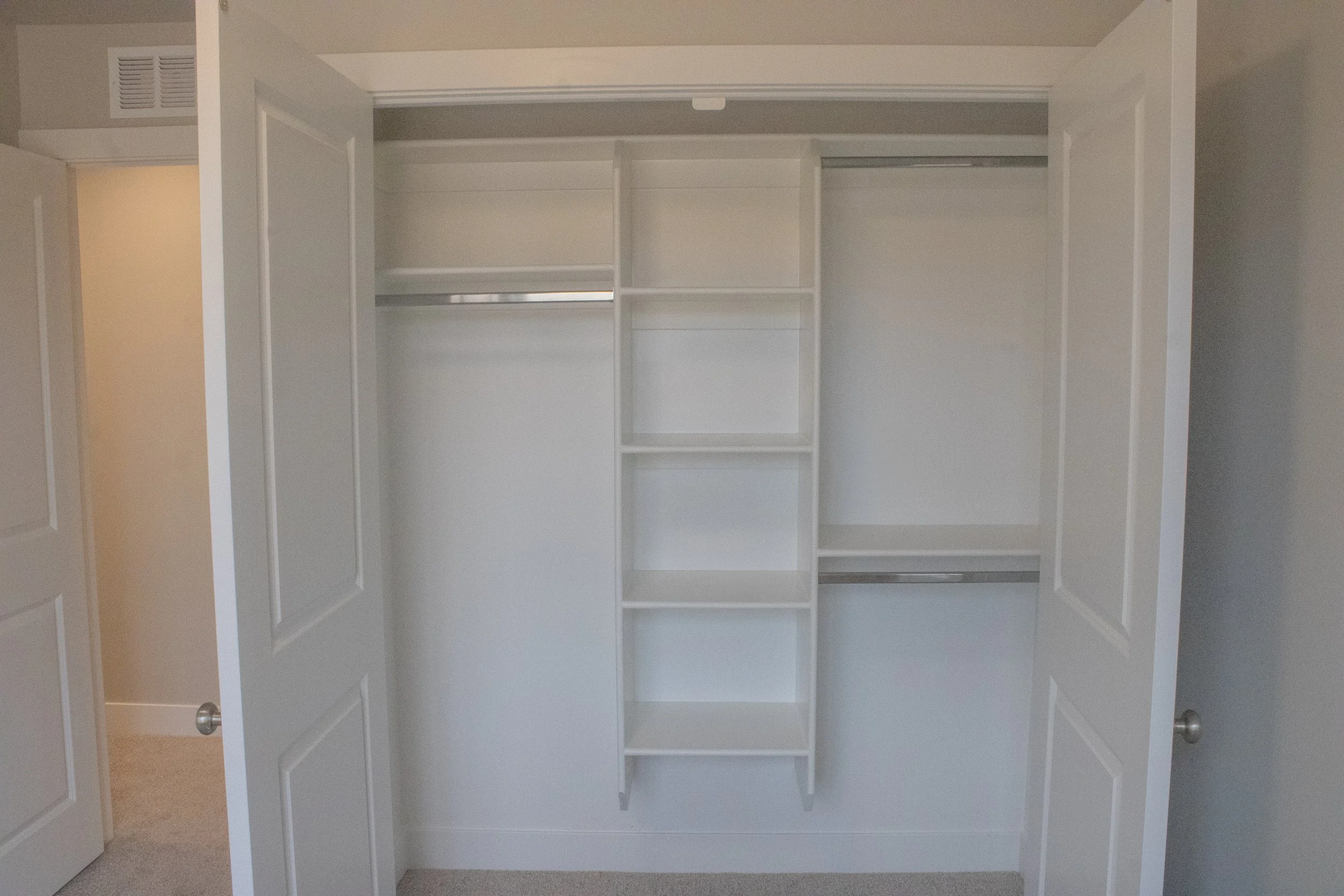 An open reach-in closet with white double doors and a built-in closet organizer. The system includes vertical shelves in the center and chrome hanging rods on the top and bottom.