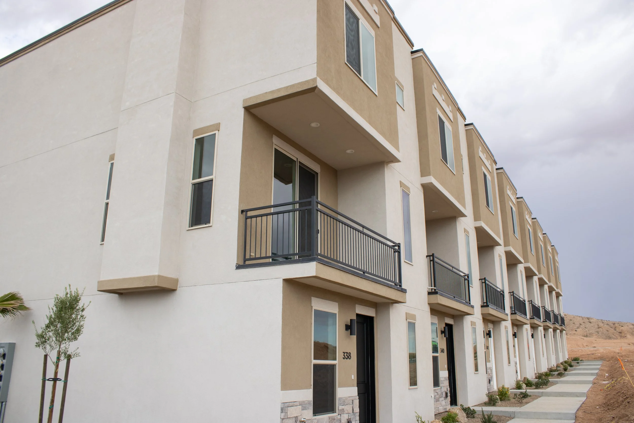 Front exterior of a modern three-story townhouse with beige and white stucco siding, a black front door, a second-floor balcony, and a stacked stone veneer base.