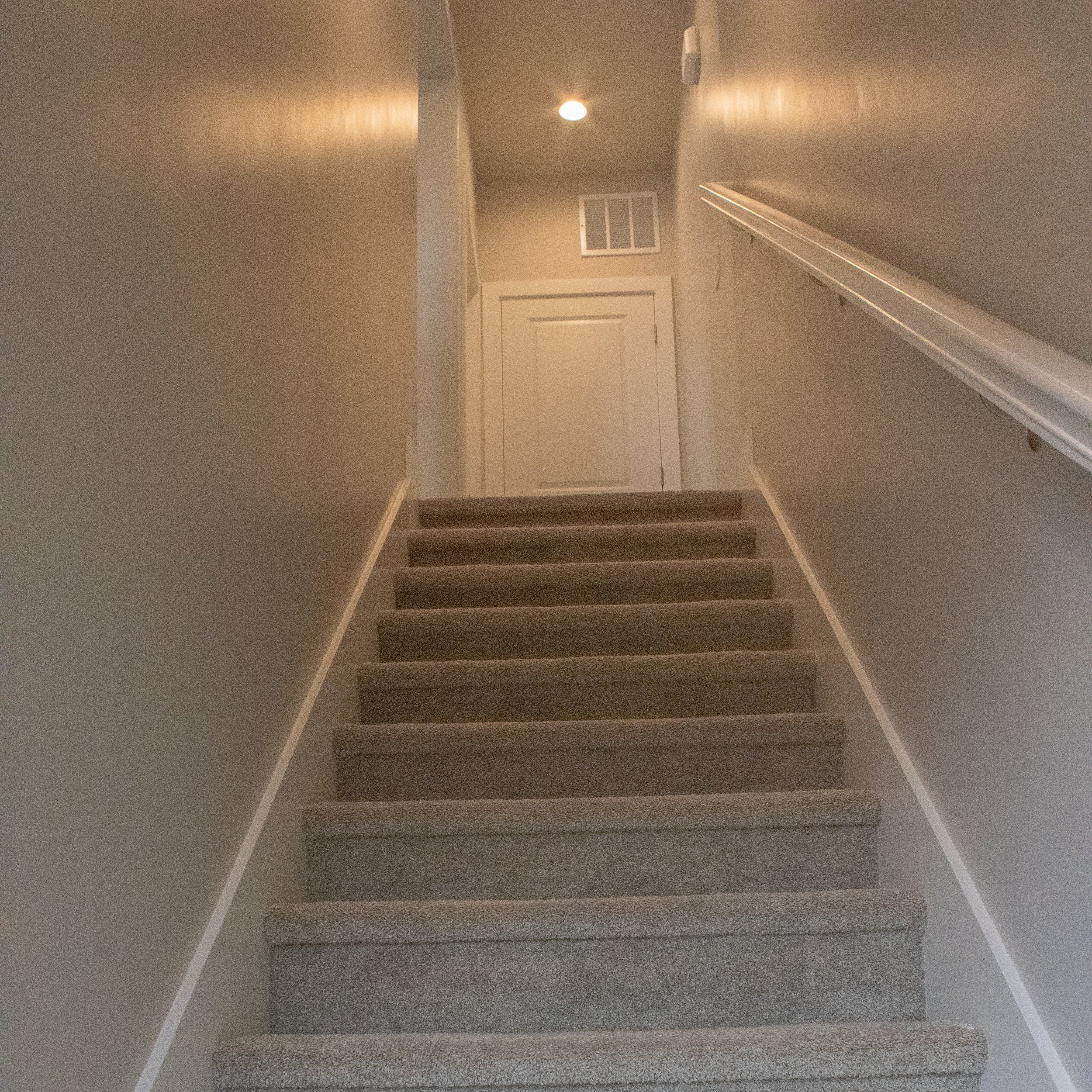 View looking up a set of interior residential stairs with light beige carpeting and white trim. The landing features a white door, a small air vent, and a recessed ceiling light.