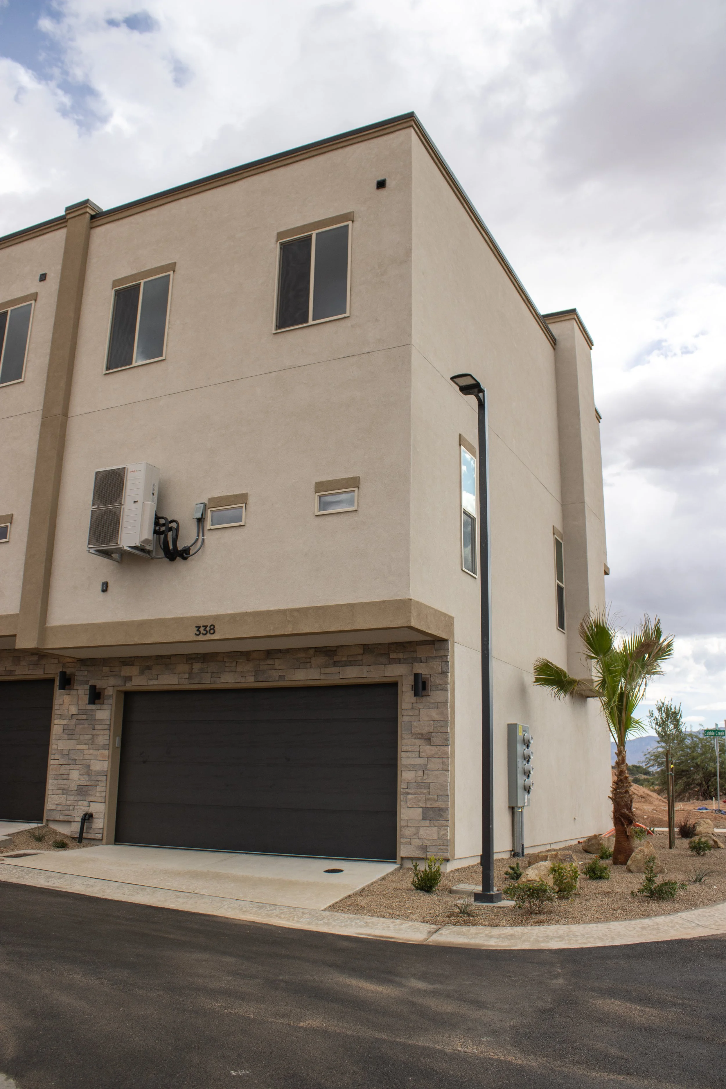 Exterior view of a row of modern townhouses with beige stucco siding, dark garage doors accented with stacked stone veneer, and exposed outdoor HVAC units.
