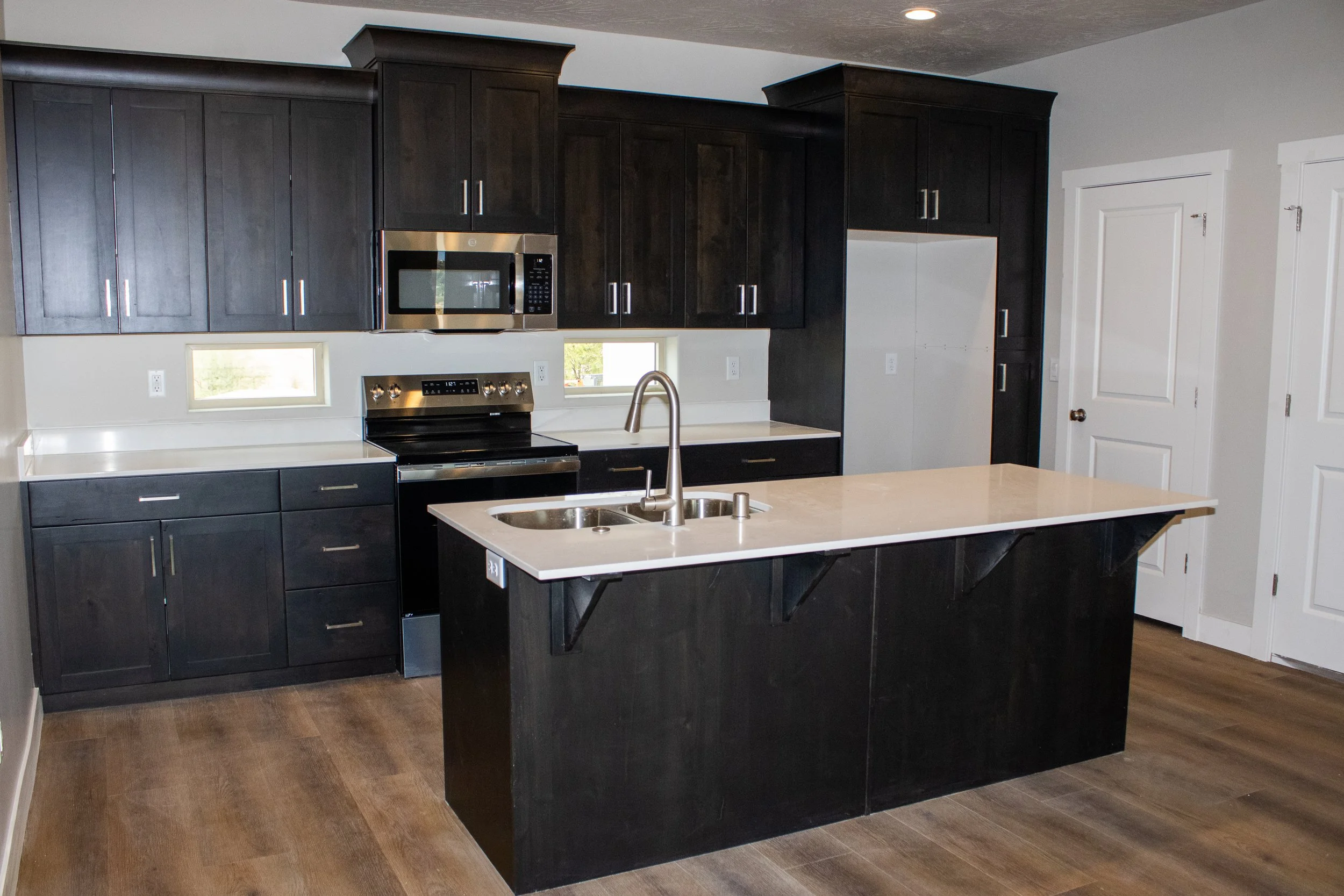 A modern kitchen featuring dark espresso shaker cabinets and a central island with a contrasting white quartz countertop. Includes stainless steel microwave and range, with wood-look flooring.