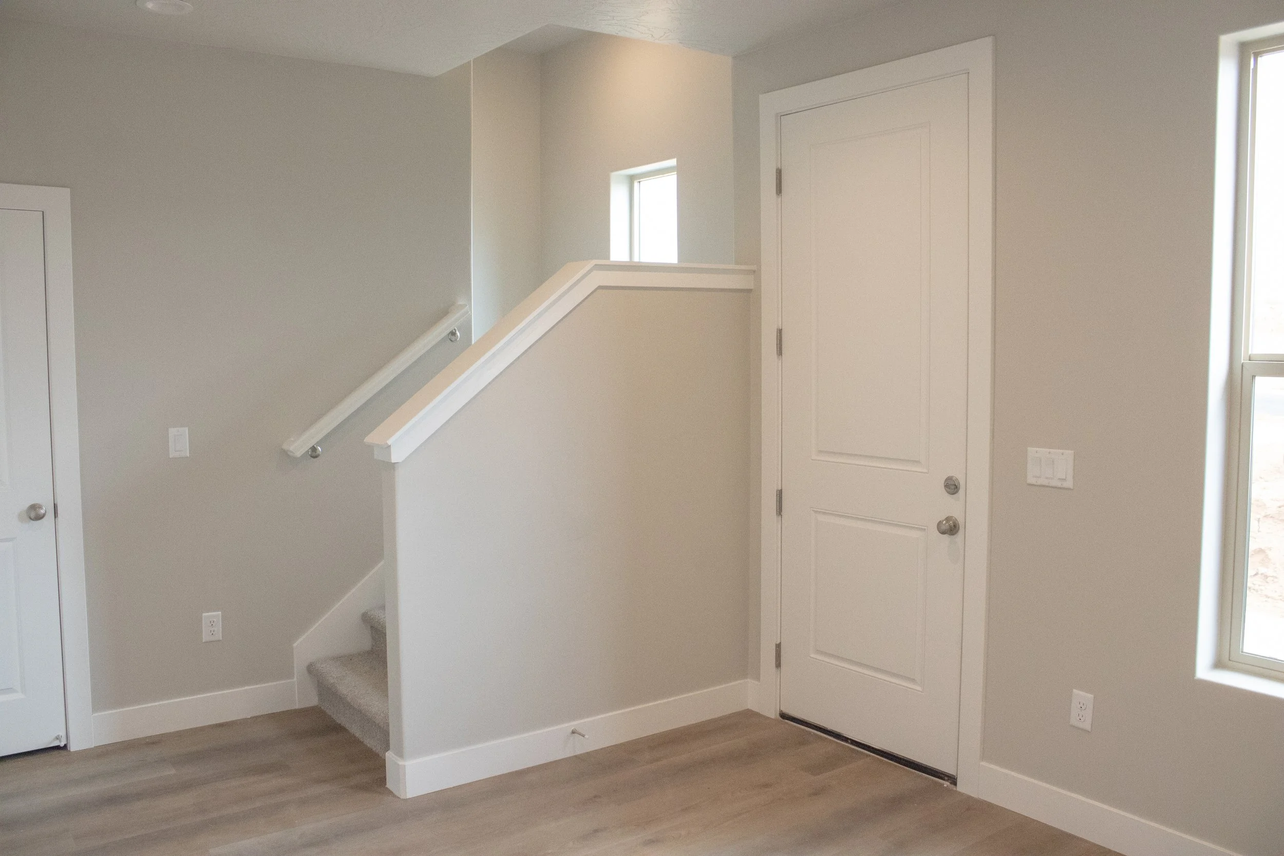 Residential entryway and staircase featuring light wood-look vinyl plank flooring, neutral paint, and crisp white trim. The photo shows the main entry door and a short flight of stairs with a white railing.
