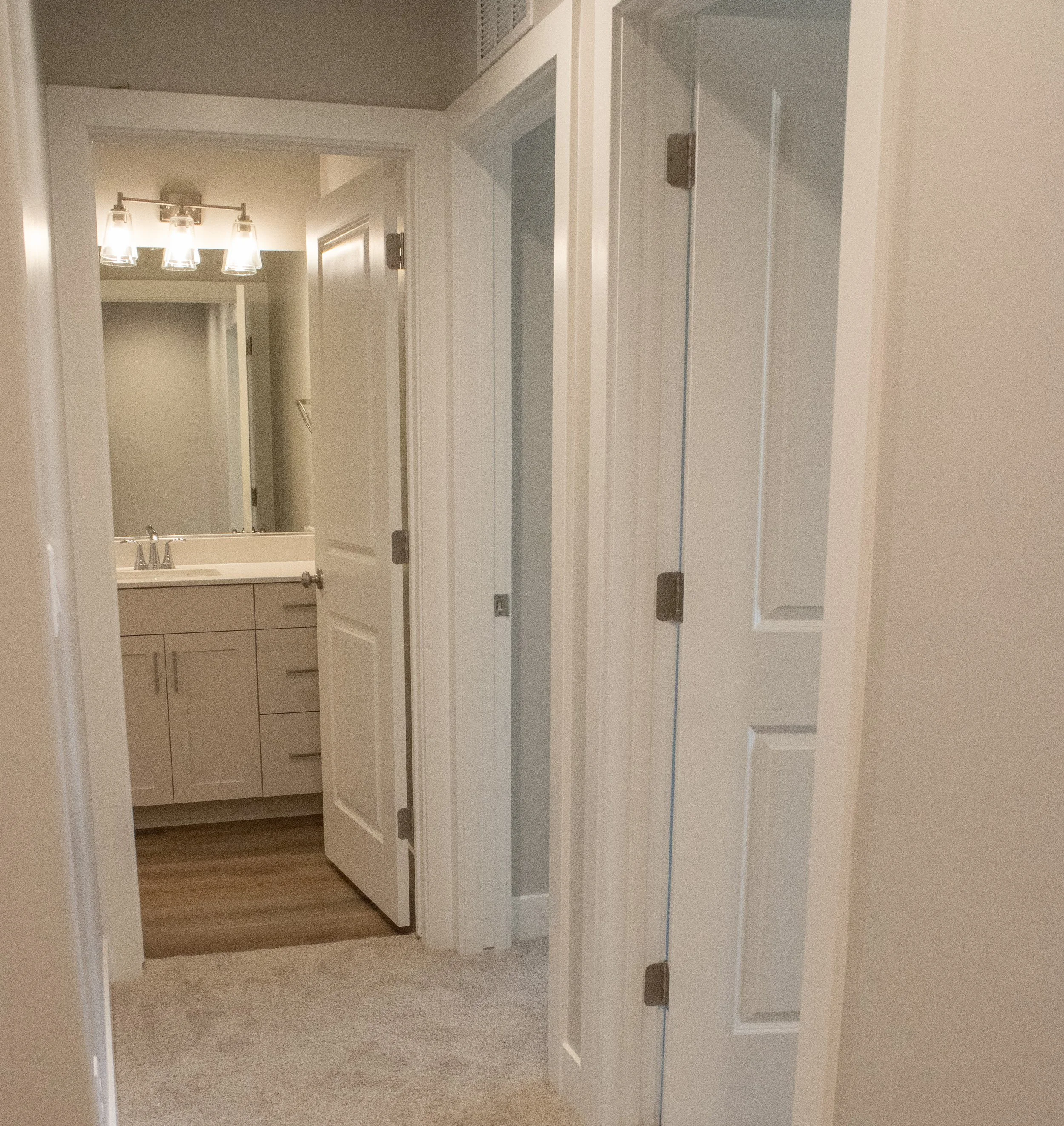 Carpeted hallway looking into an open bathroom doorway. The image features white doors, a light gray vanity, and a mirror with a four-light vanity fixture.