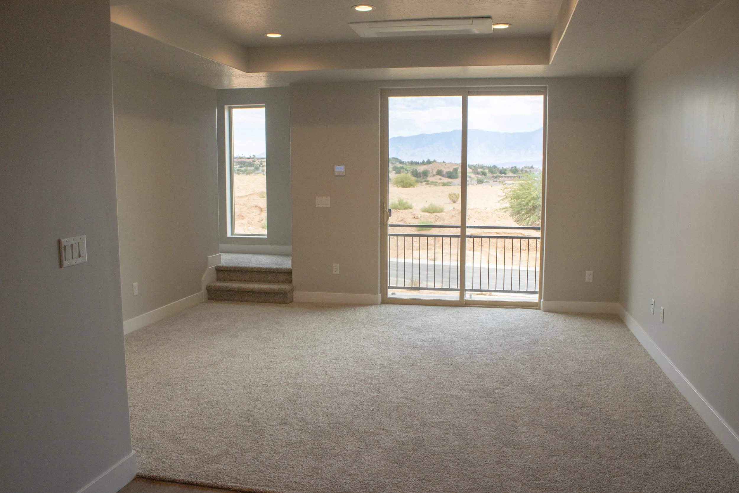 An empty bedroom in a new construction home featuring neutral gray walls, light beige carpet, white baseboard trim, and a modern ceiling fan. A large window offers a view of the surrounding landscape.