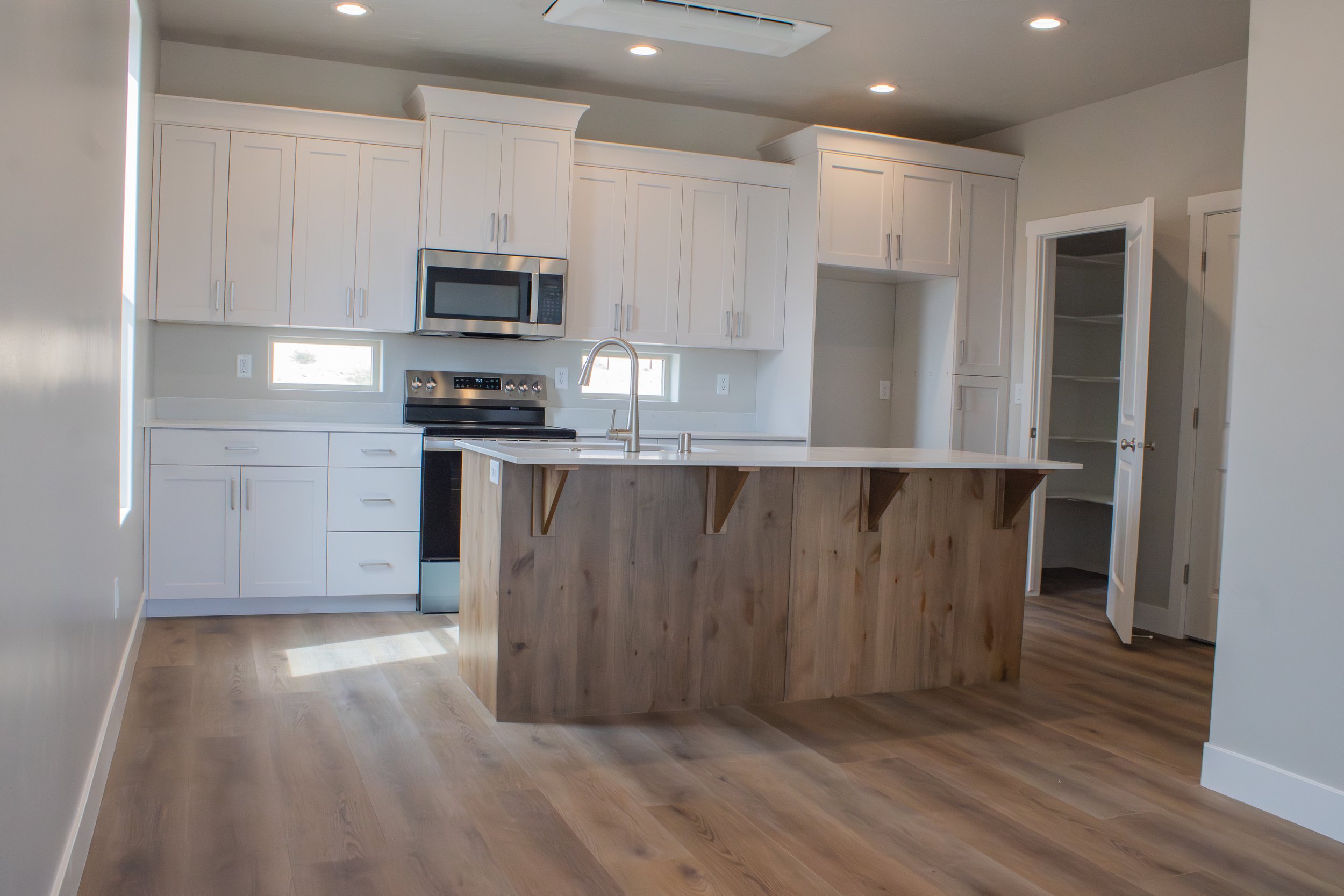 A bright, modern kitchen featuring white shaker cabinets and a central island clad in natural wood grain. The kitchen has a white countertop, stainless steel appliances.