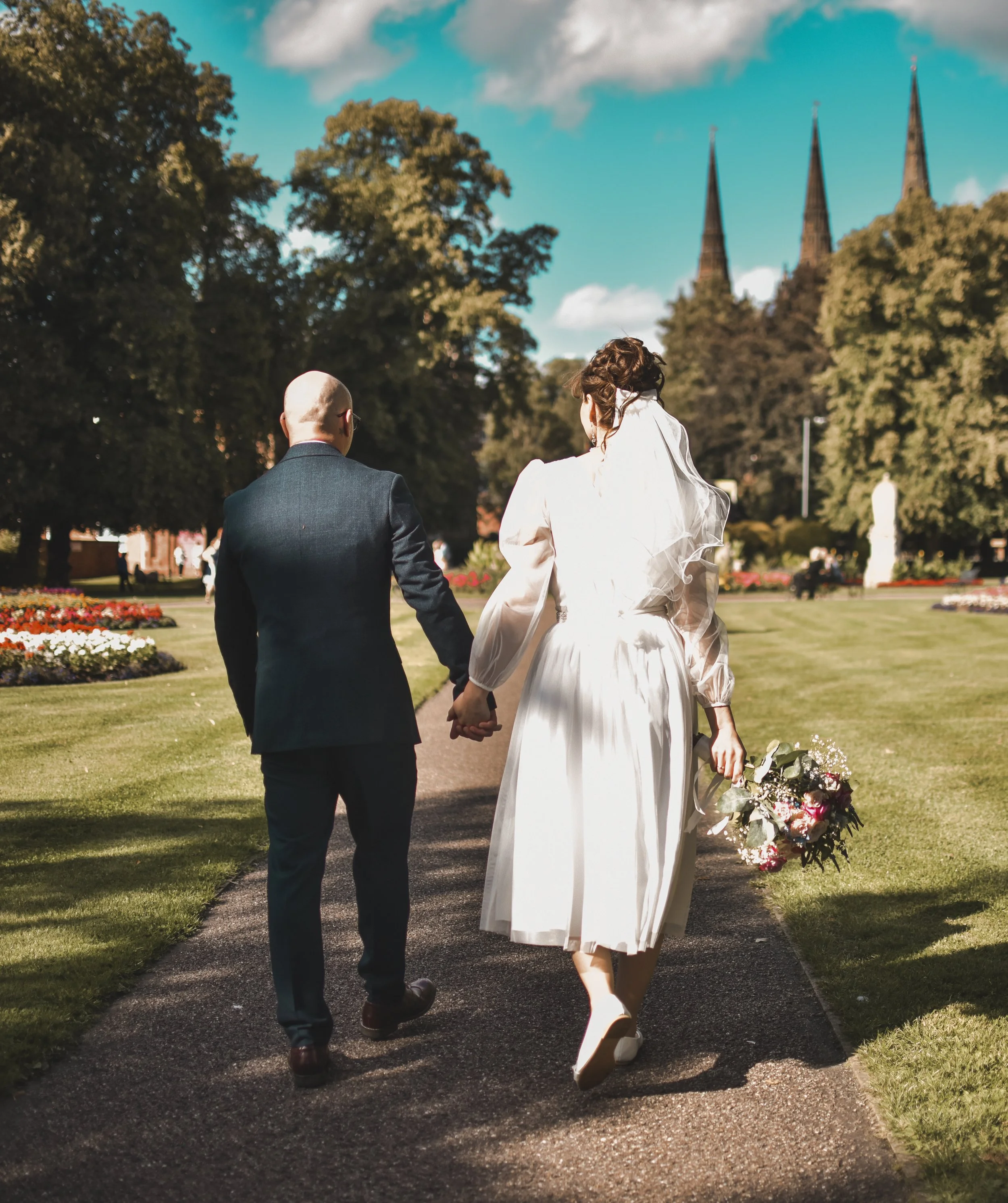 A bride and groom walking hand in hand outdoors on a sunny day, with a church with three tall steeples visible in the background and surrounded by trees.