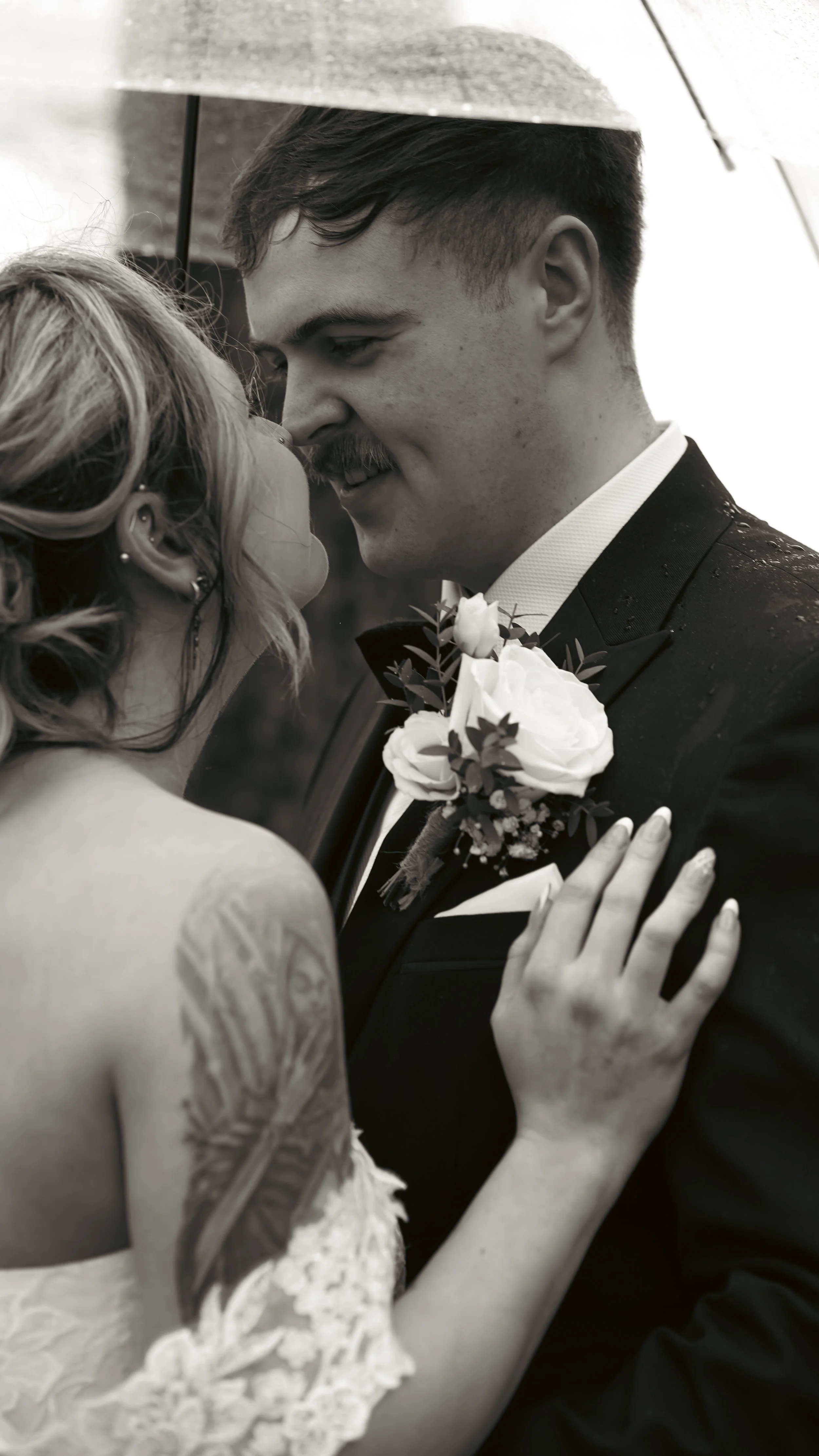 Black and white photo of a bride and groom close, smiling, under an umbrella, about to kiss during their wedding.