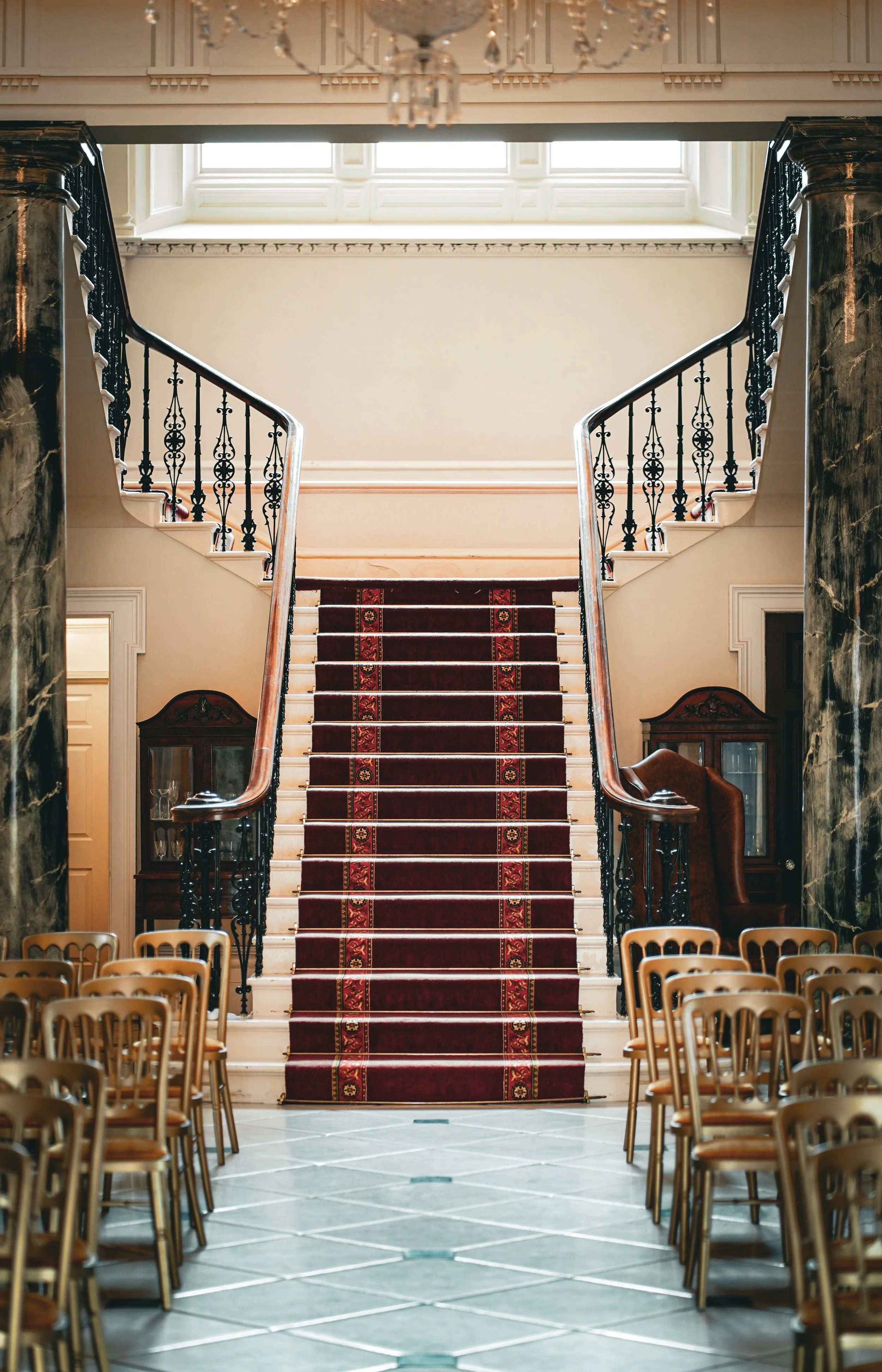 Elegant interior with grand staircase, red carpet runner, and wooden chairs arranged on either side, in a spacious, well-lit room.
